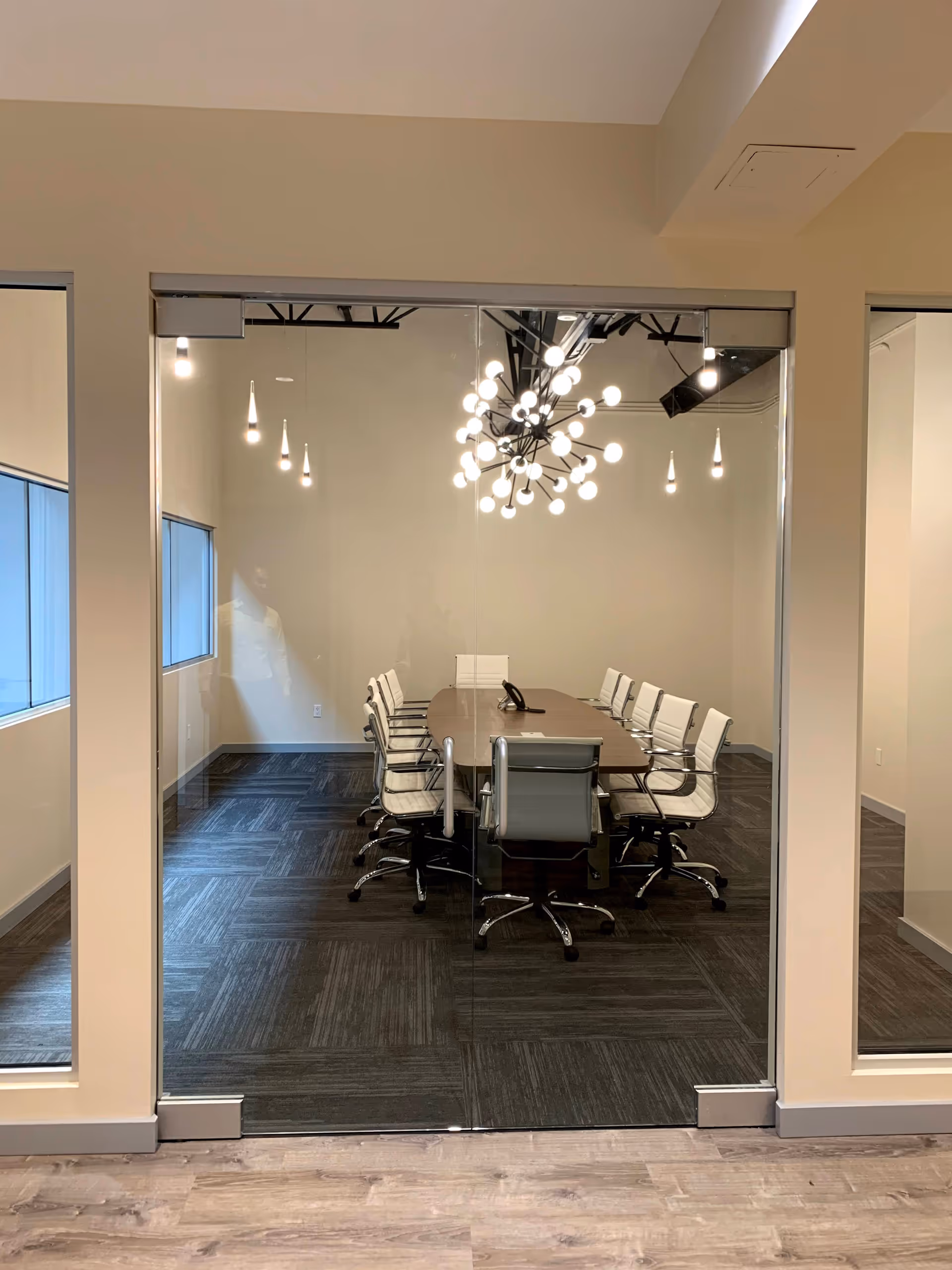 Glass-walled conference room with a long table, white chairs, and a modern chandelier.