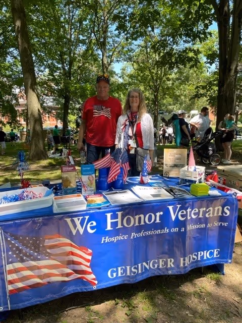 Two adults stand behind a blue We Honor Veterans table at an outdoor park event.