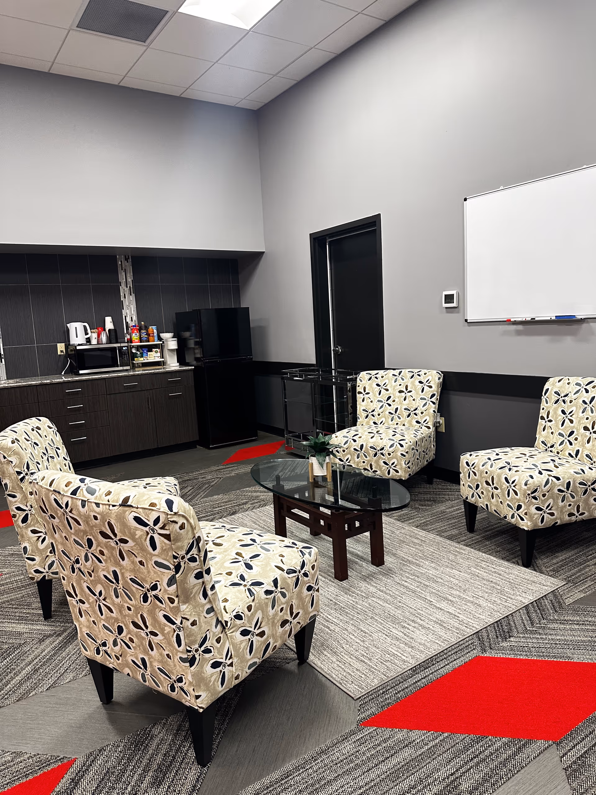 A lounge area with patterned chairs around a glass coffee table and kitchenette in back.