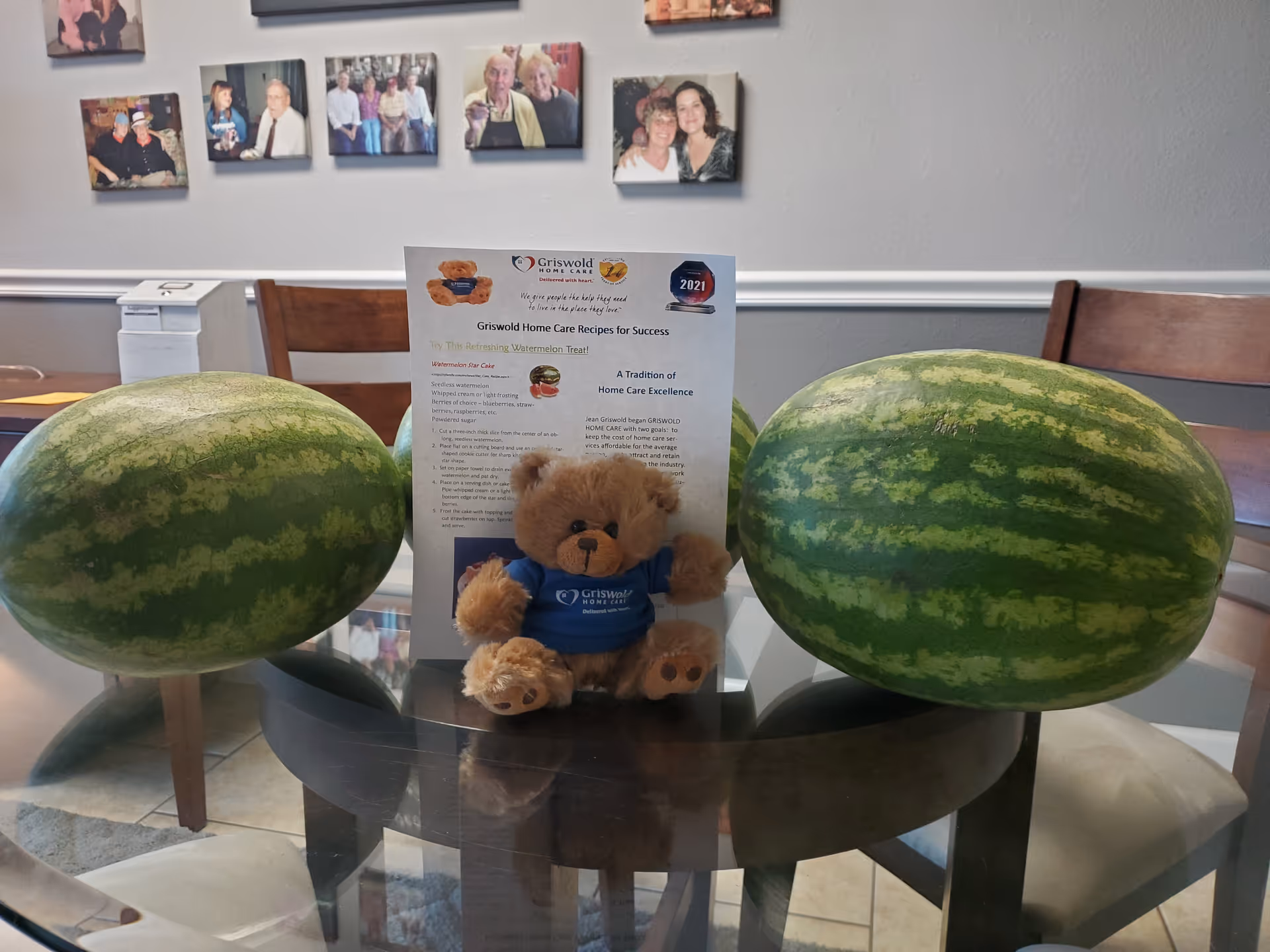 Two large watermelons flank a teddy bear and a flyer on a glass table.