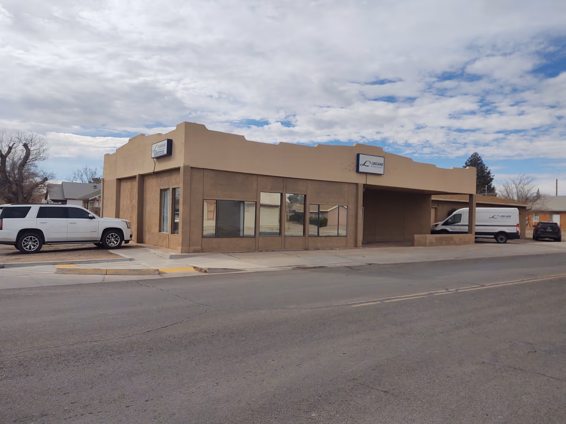 Tan Lincare building with parked white vehicles along a street under a partly cloudy sky.