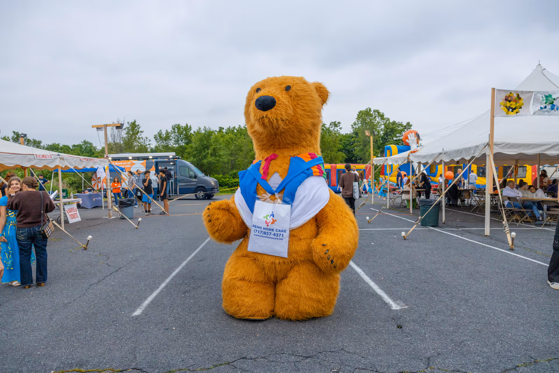 Large orange teddy bear mascot in blue vest stands outdoors at a festival with tents nearby.