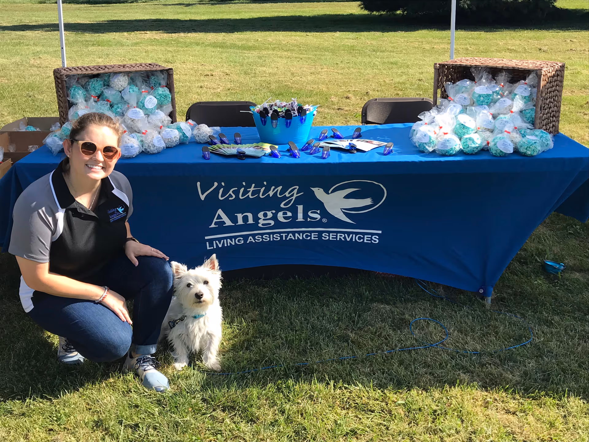 Smiling woman beside a table with Visiting Angels banner at an outdoor event with a dog