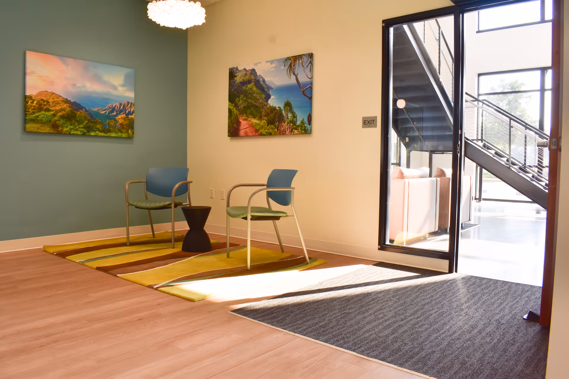 Bright interior lounge with two chairs, colorful rugs, wall art, and a glass door.
