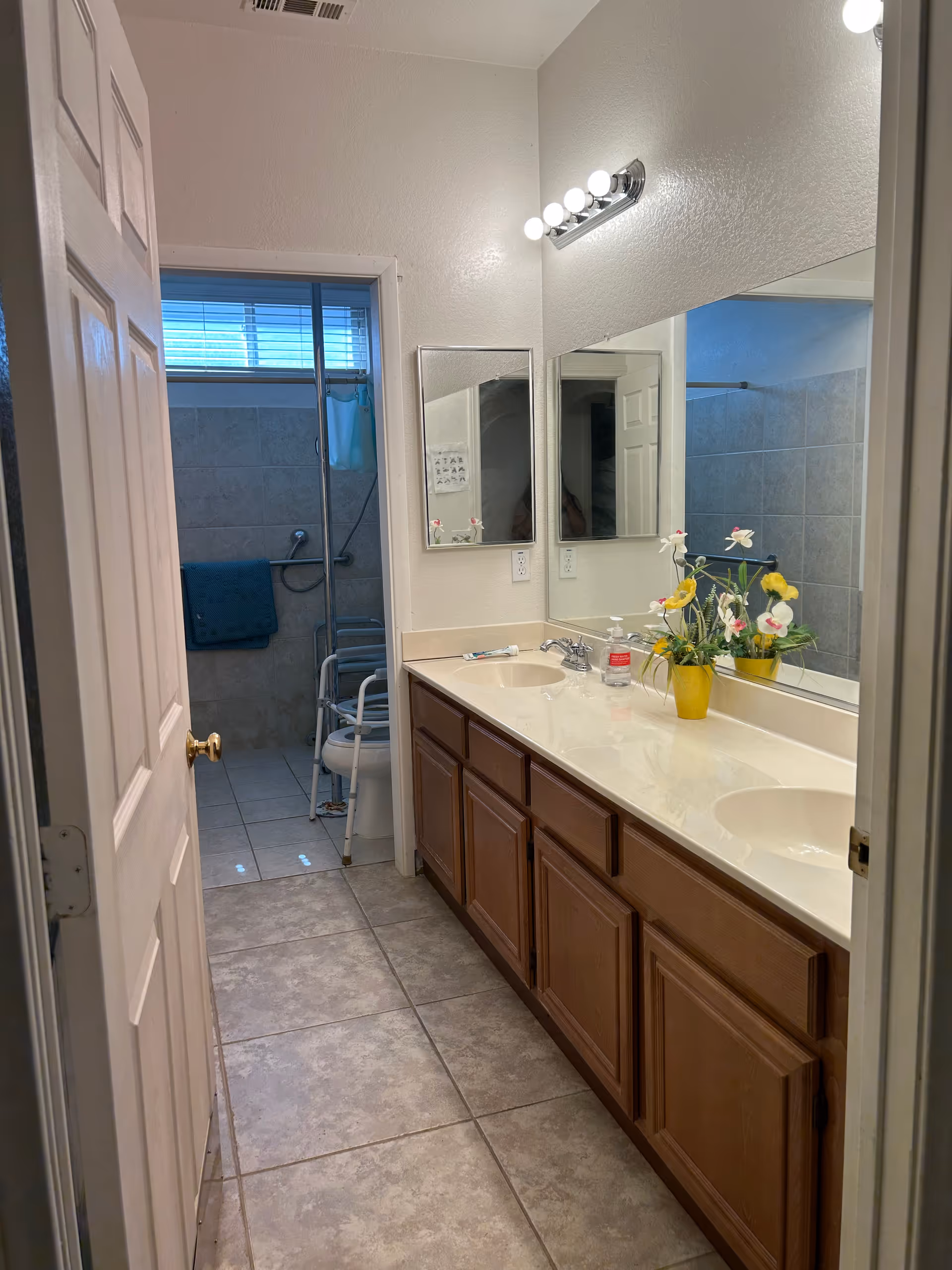 Bathroom features a double-sink vanity, mirrors, wooden cabinets, and a shower in the back.