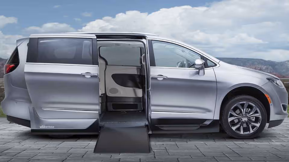 Silver wheelchair-accessible van with extended ramp parked on a stone driveway.
