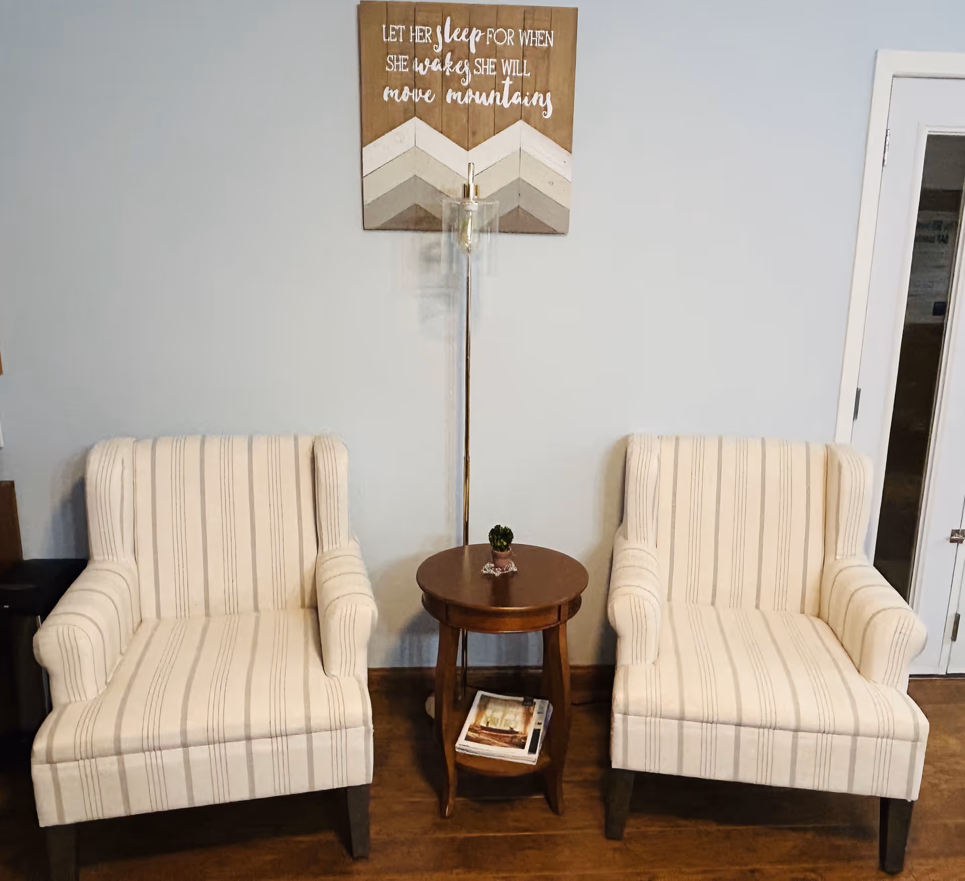 Two striped armchairs flank a round wooden table with magazines beneath, against a light blue wall.