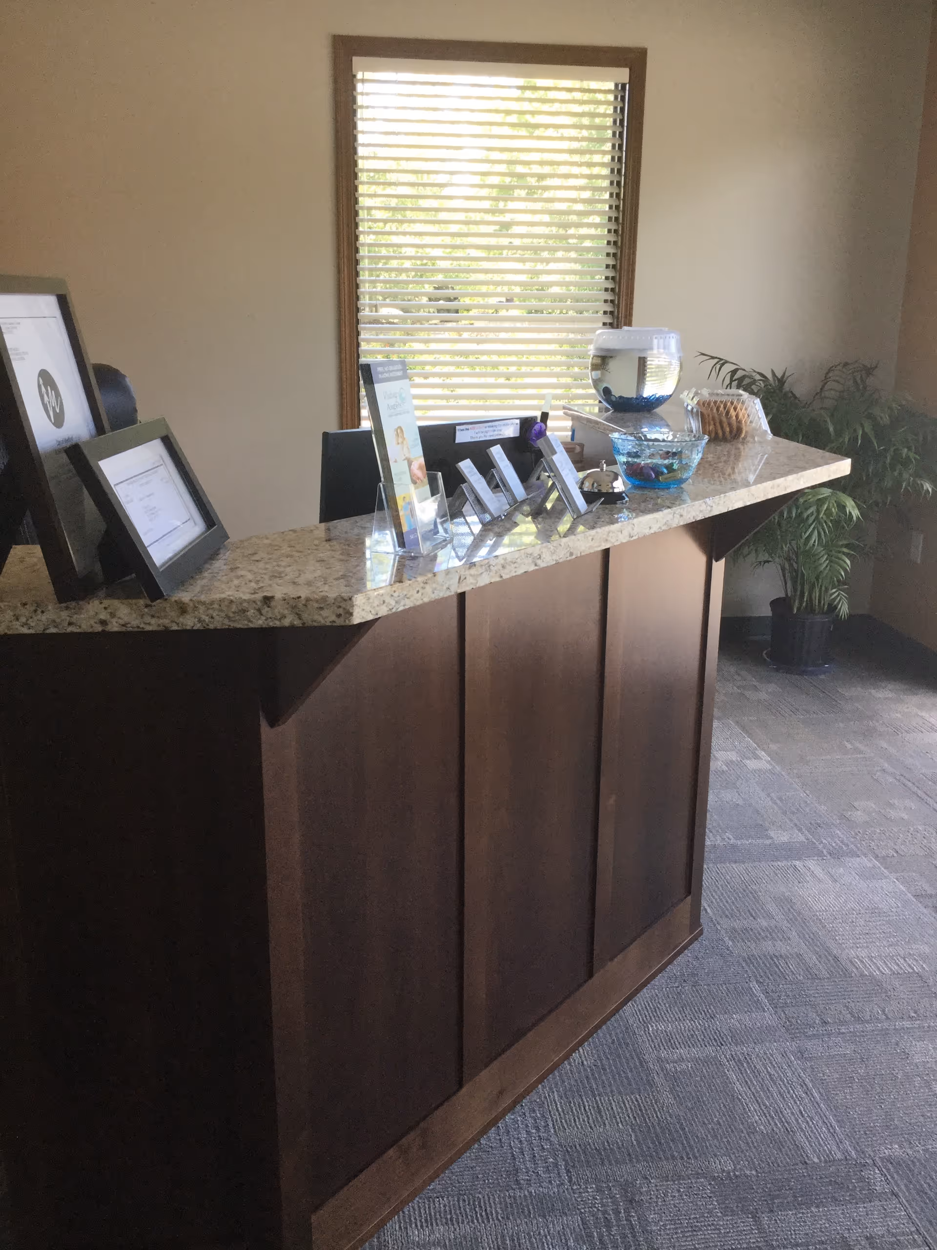 Reception counter in a lobby with framed notices, beverage station, and window blinds.