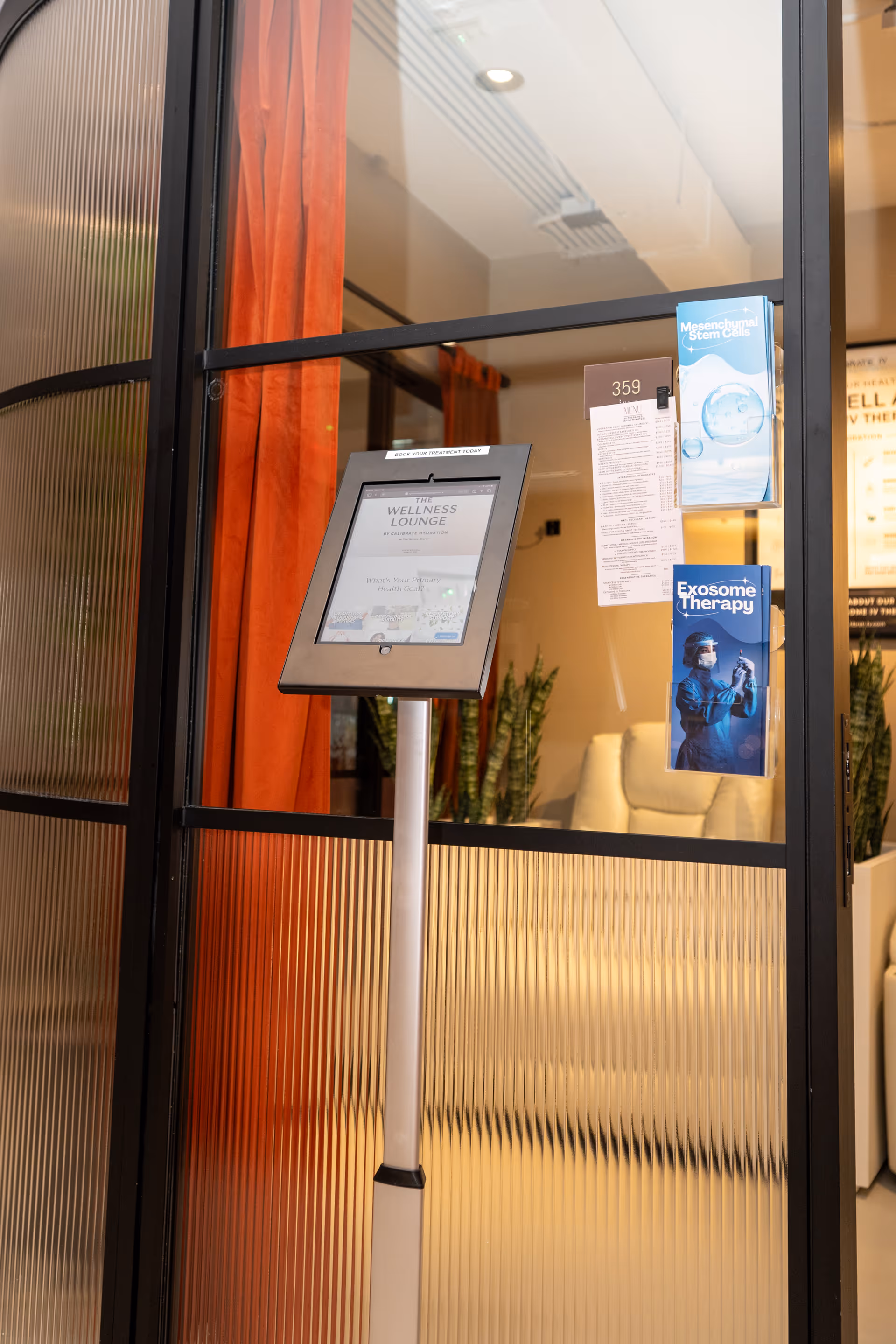 Touchscreen information kiosk in a glass-walled lobby with posters and plants.