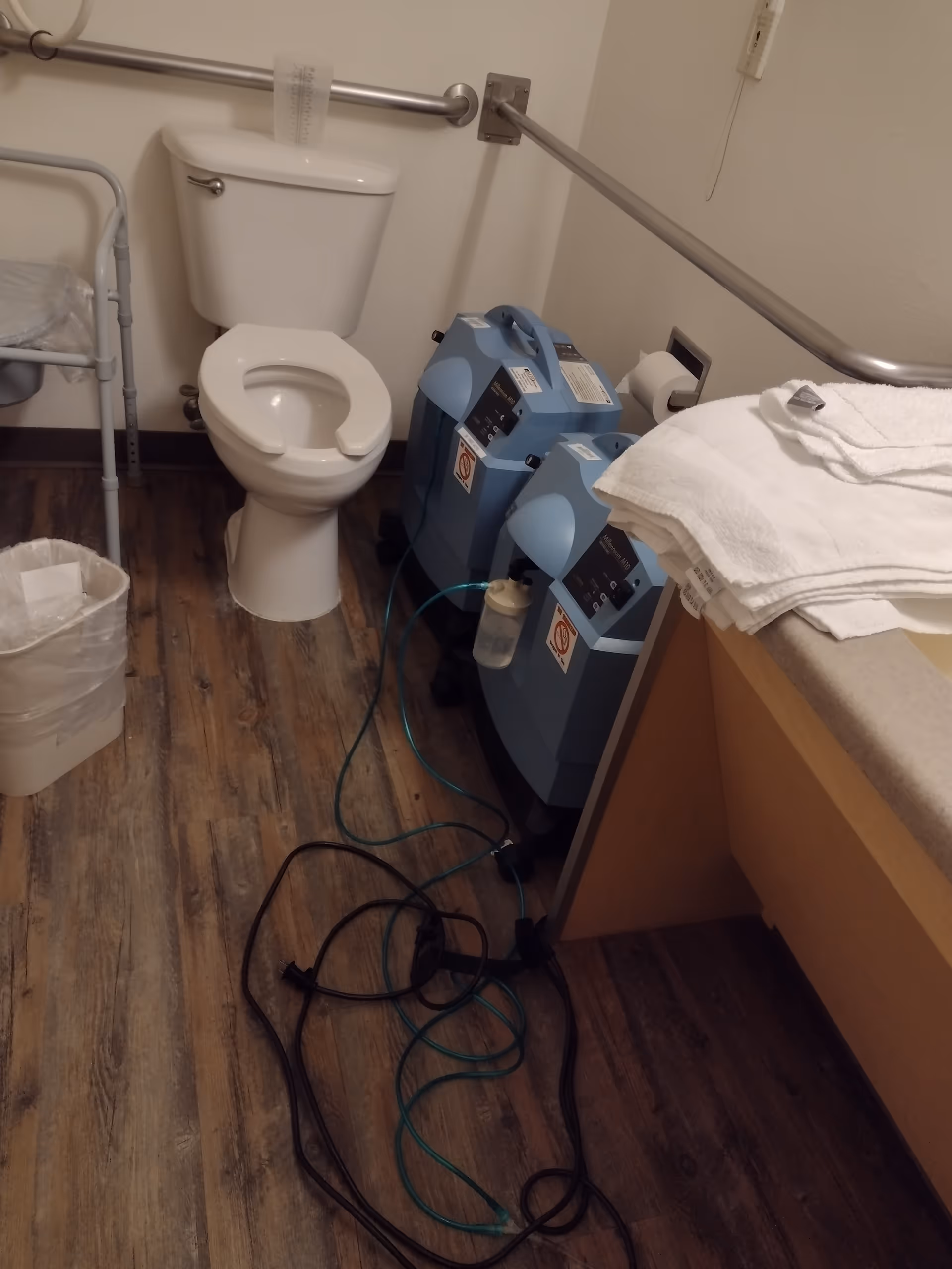 Small bathroom with toilet, medical machines, towels, and tangled cords on wooden floor.