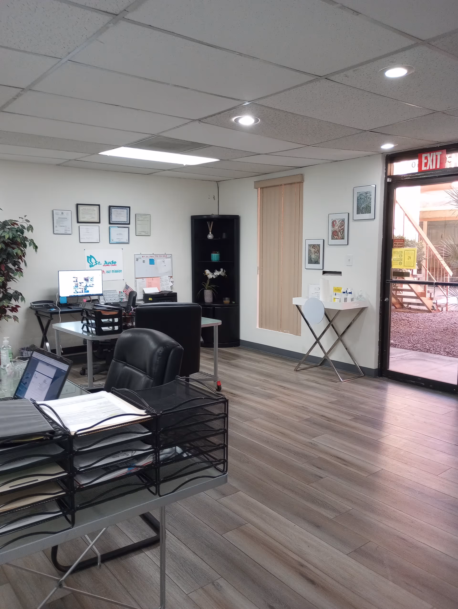 Office reception area with desk, computer, filing trays, certificates, and exit door.
