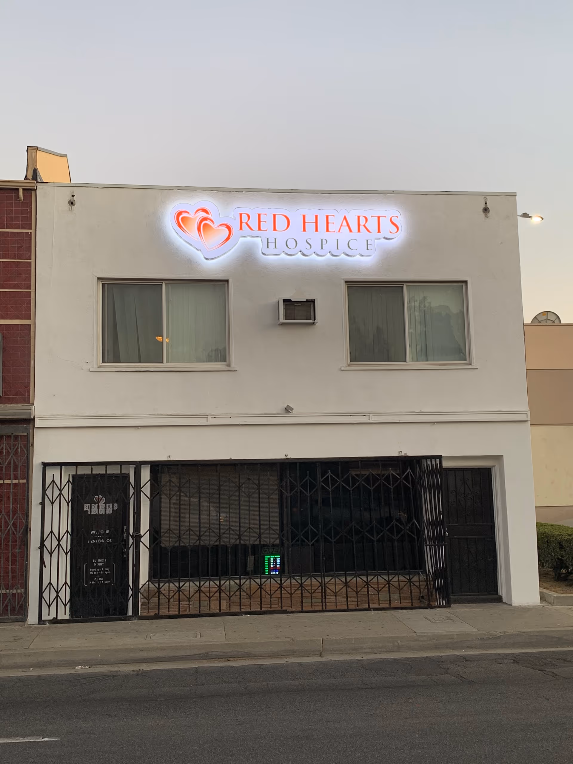Two-story white building with an illuminated Red Hearts Hospice sign and a metal gate.