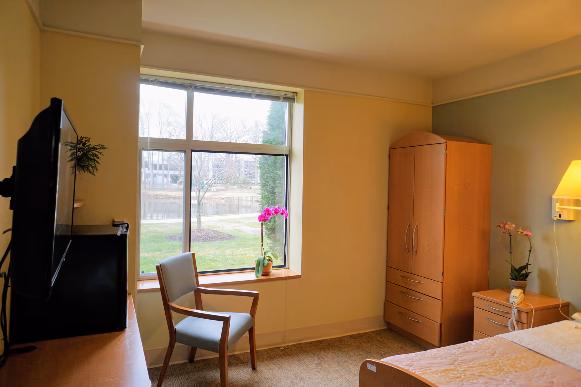 Senior living facility bedroom with chair by window, wardrobe, dresser, bed, and orchids.