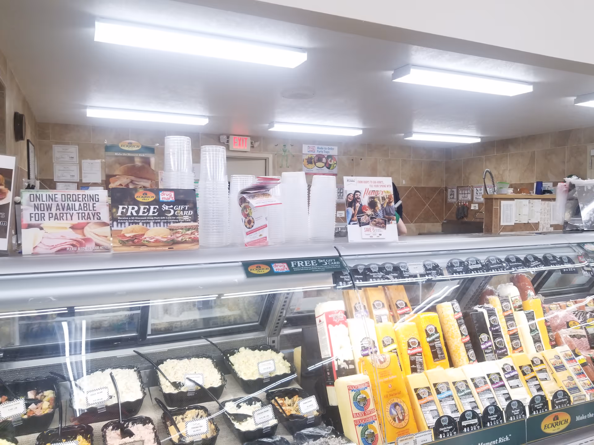 Inside Discount Drug Mart, a refrigerated deli counter displays meats, cheeses, and stacked cups.