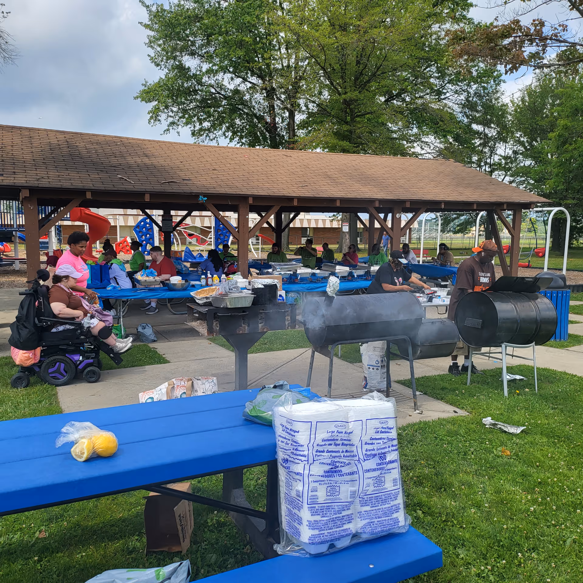 An outdoor pavilion hosts a community cookout with tables, grills, and residents.
