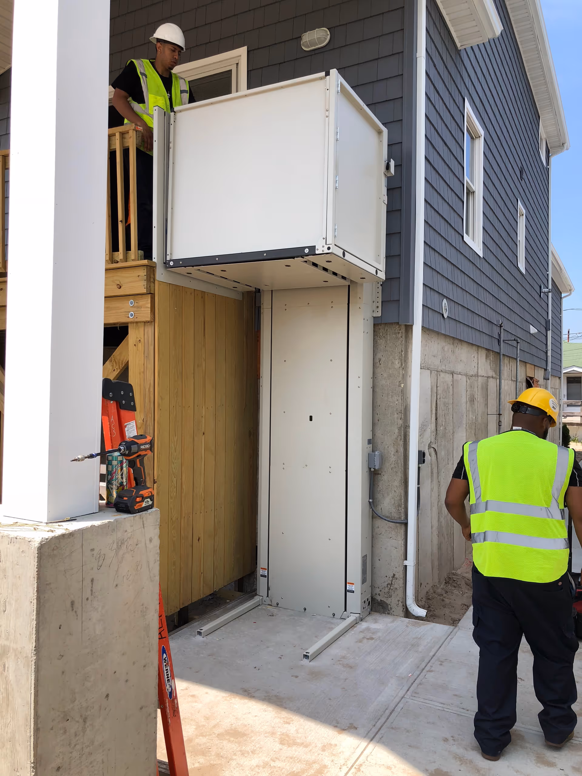 Two construction workers in safety vests install a lift on a building exterior.