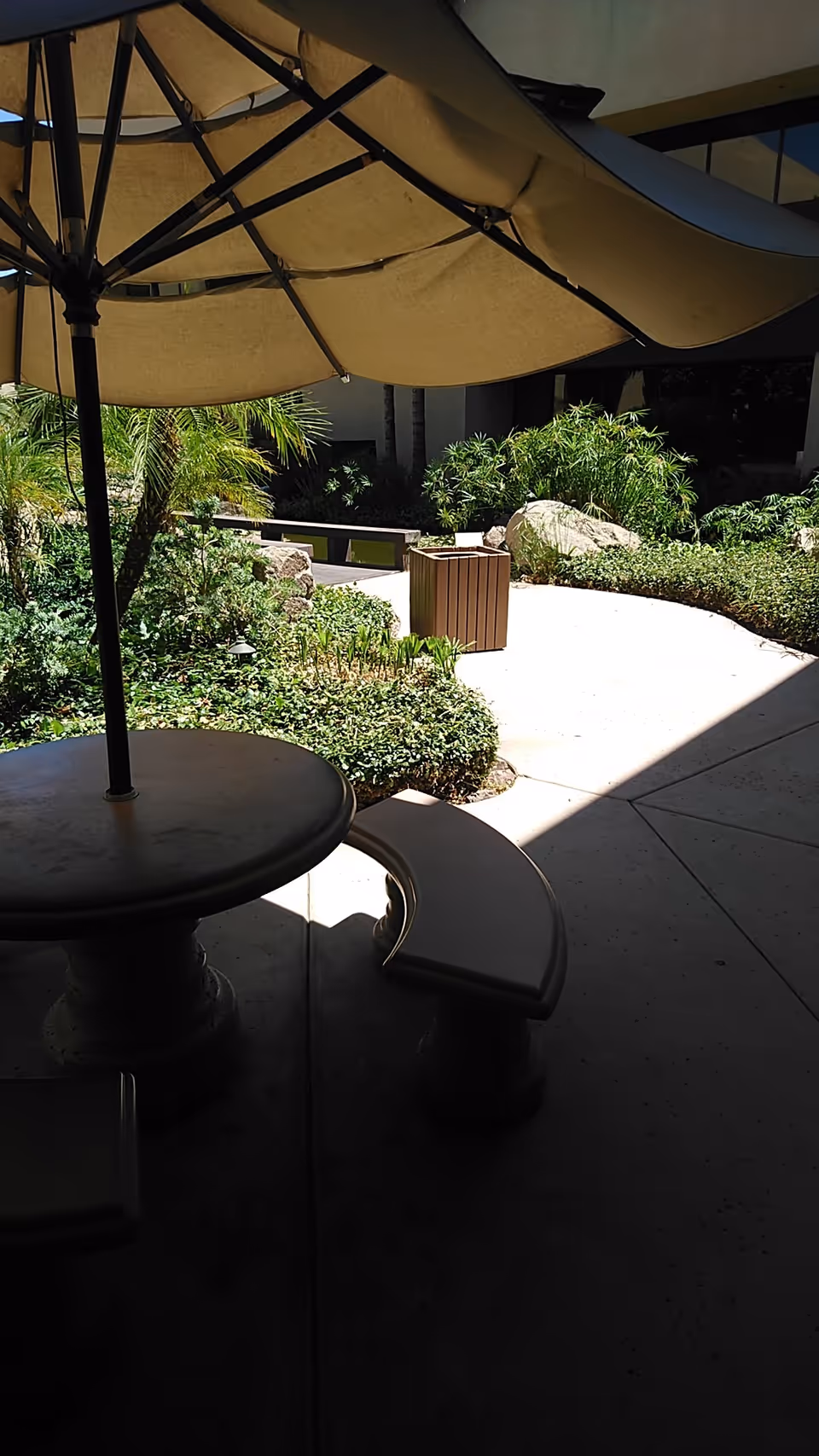 Outdoor courtyard with umbrella-covered seating and lush plants near a trash bin.
