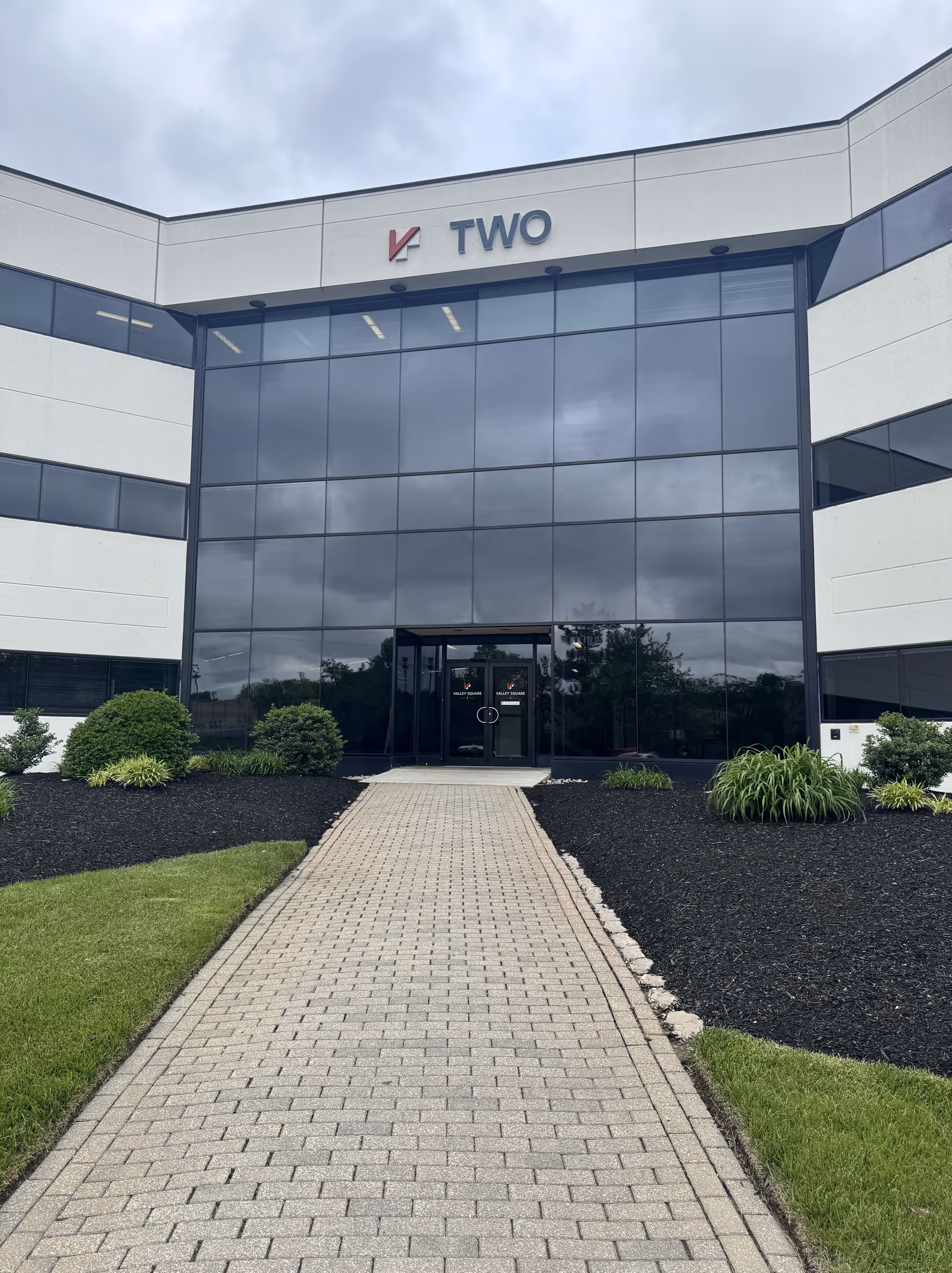 Modern glass-front building entrance with a paved walkway and surrounding landscaping.