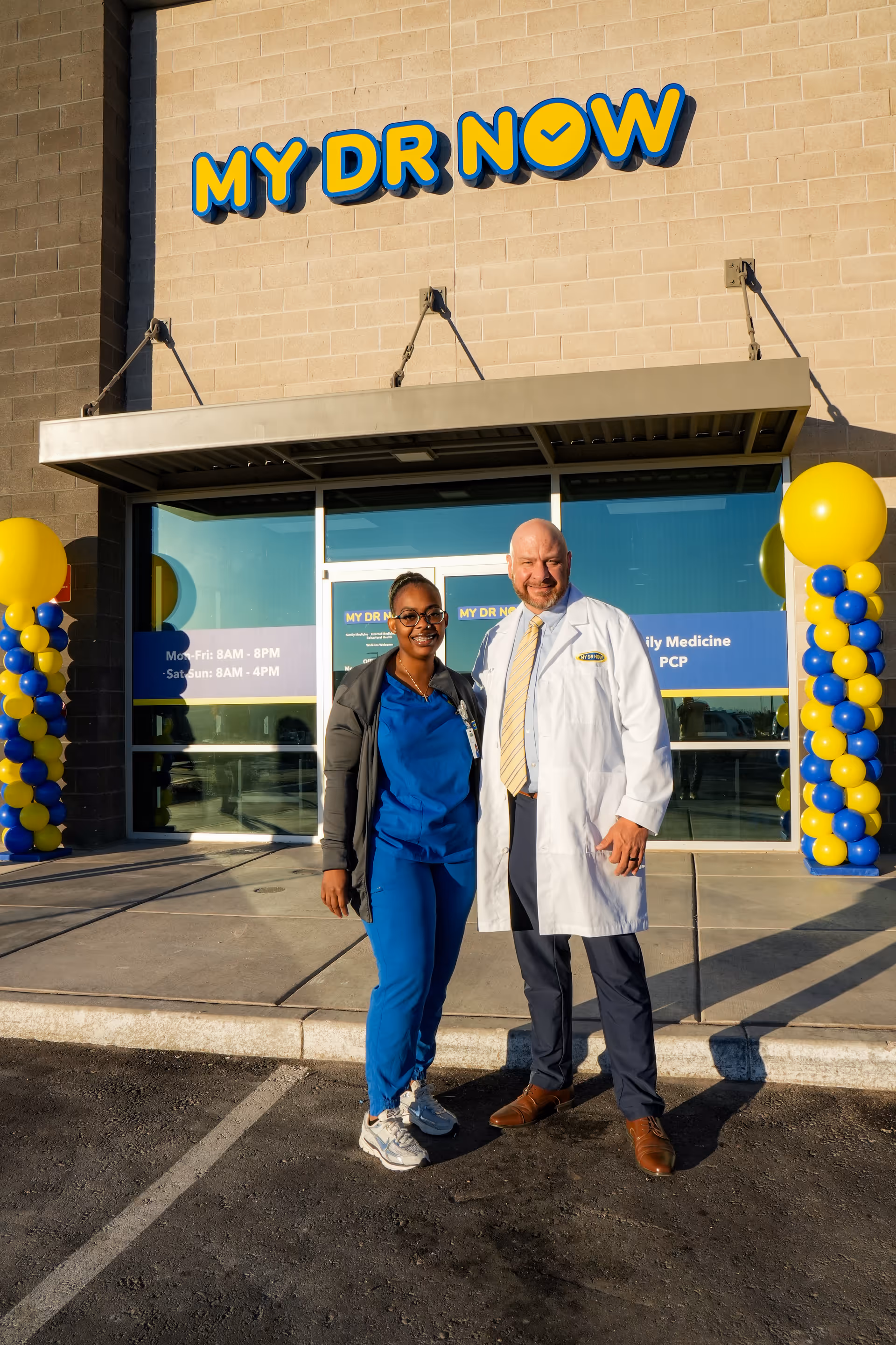 Two people in medical attire stand outside the MY DR NOW entrance with yellow balloons.