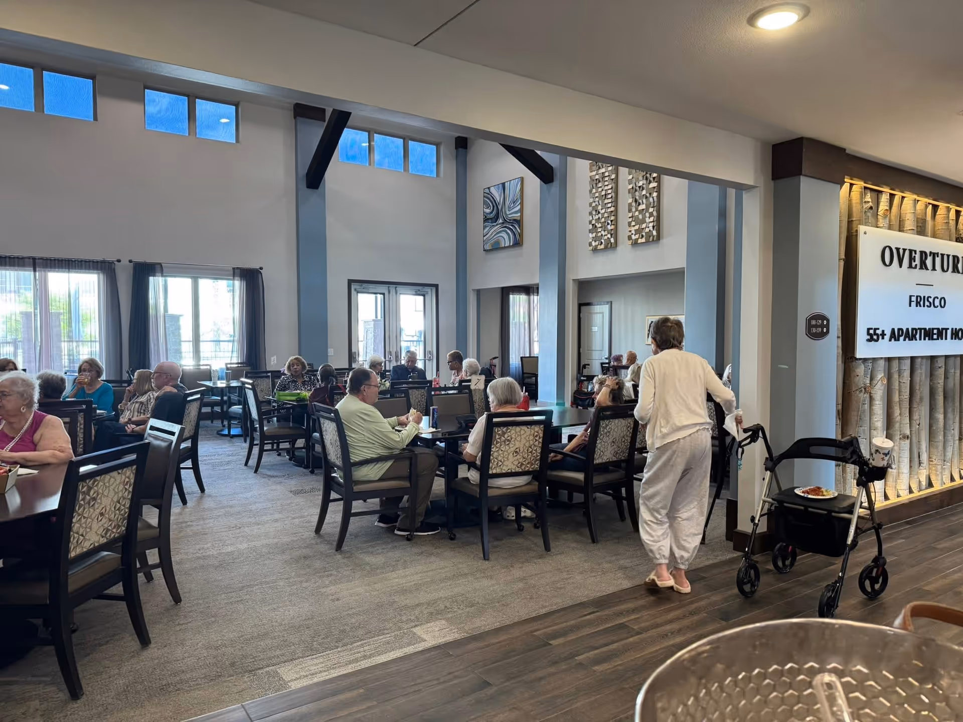 Senior residents are seated at tables in a bright dining room, with a caregiver nearby.