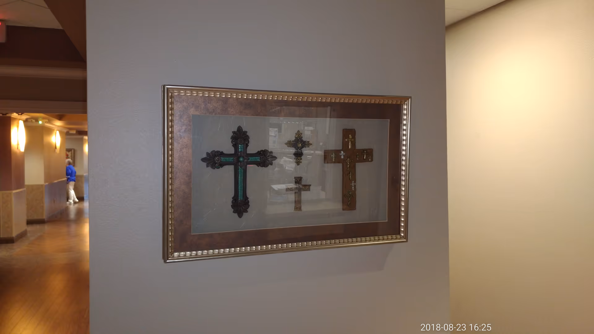 Hallway interior featuring framed cross artwork on a gray wall, with a person visible in distance.