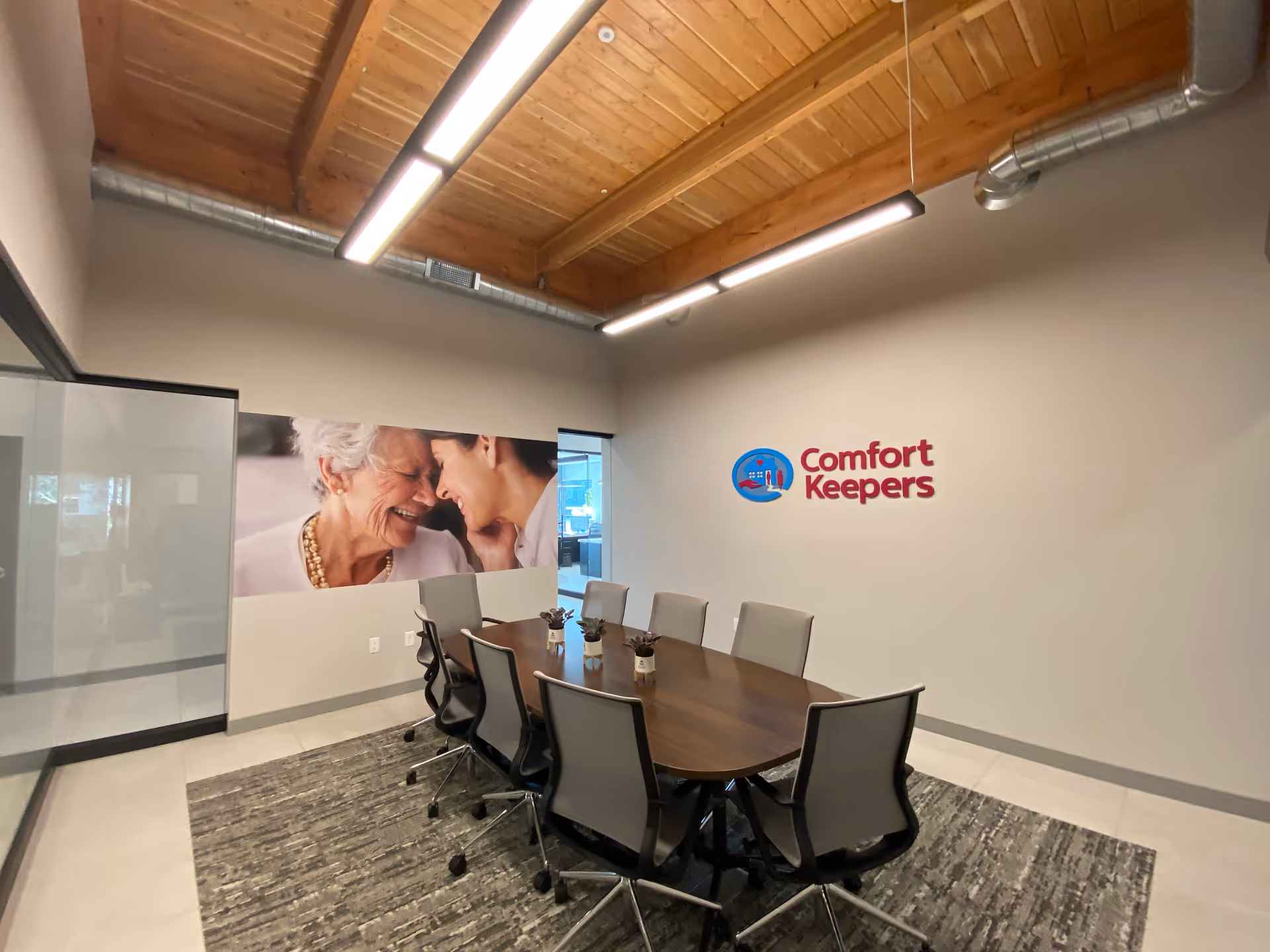 Conference room with wooden ceiling beams, long table, gray chairs, Comfort Keepers logo on wall.