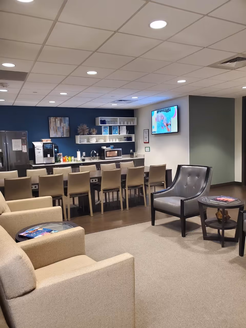 Interior senior living lounge with beige seating, a long counter, shelves, and a wall TV.