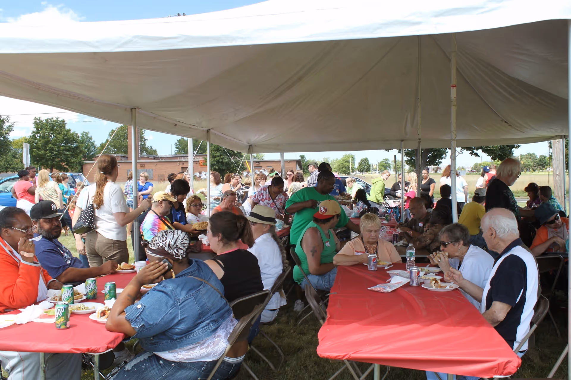 A large outdoor tent hosts a community meal with many seniors seated at red tables.