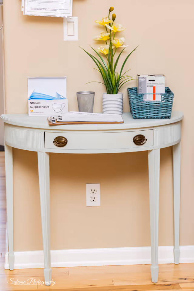 A cream console table with a vase of yellow flowers, masks, and papers against beige wall.