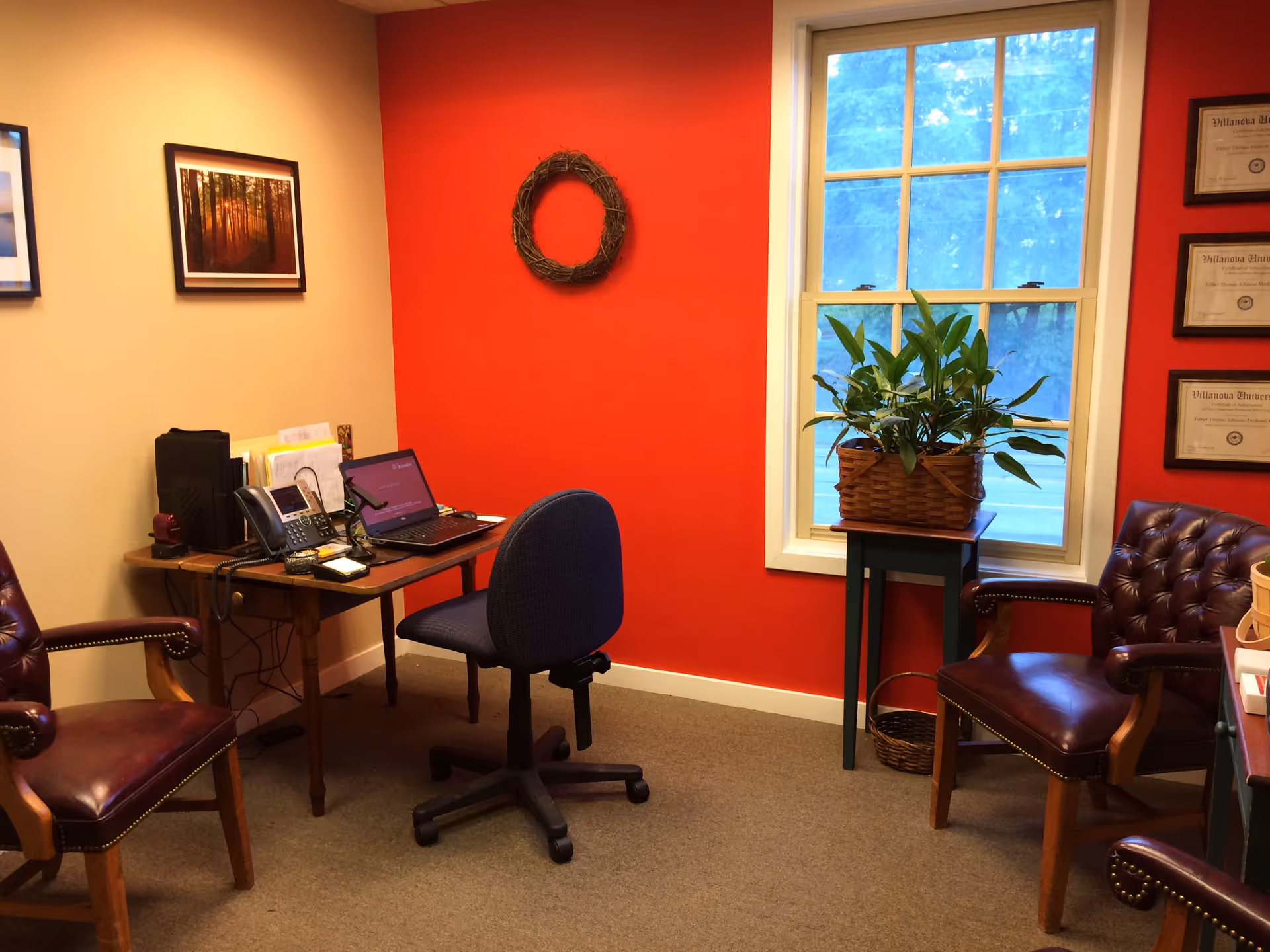 Office reception area with red accent wall, desk, laptop, and potted plant by window.