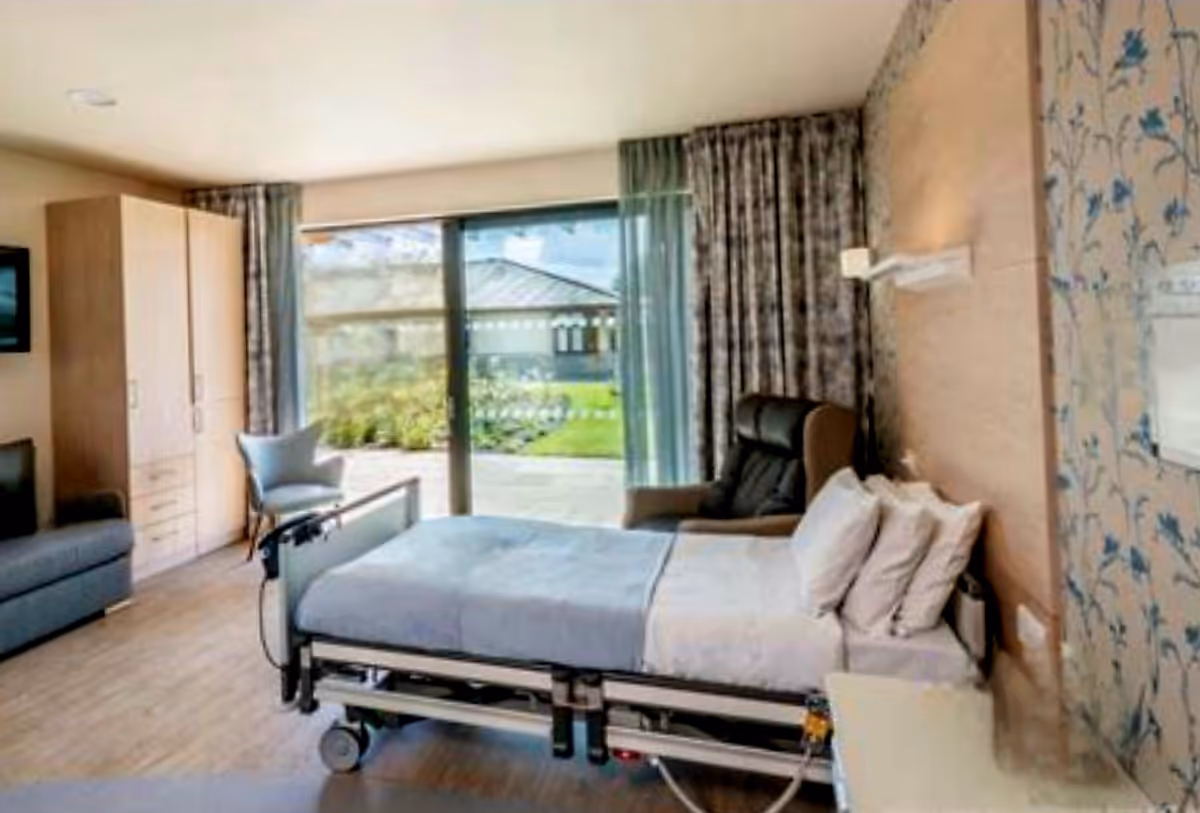Hospital-style patient room with adjustable bed, wardrobe, seating, and a large window view.
