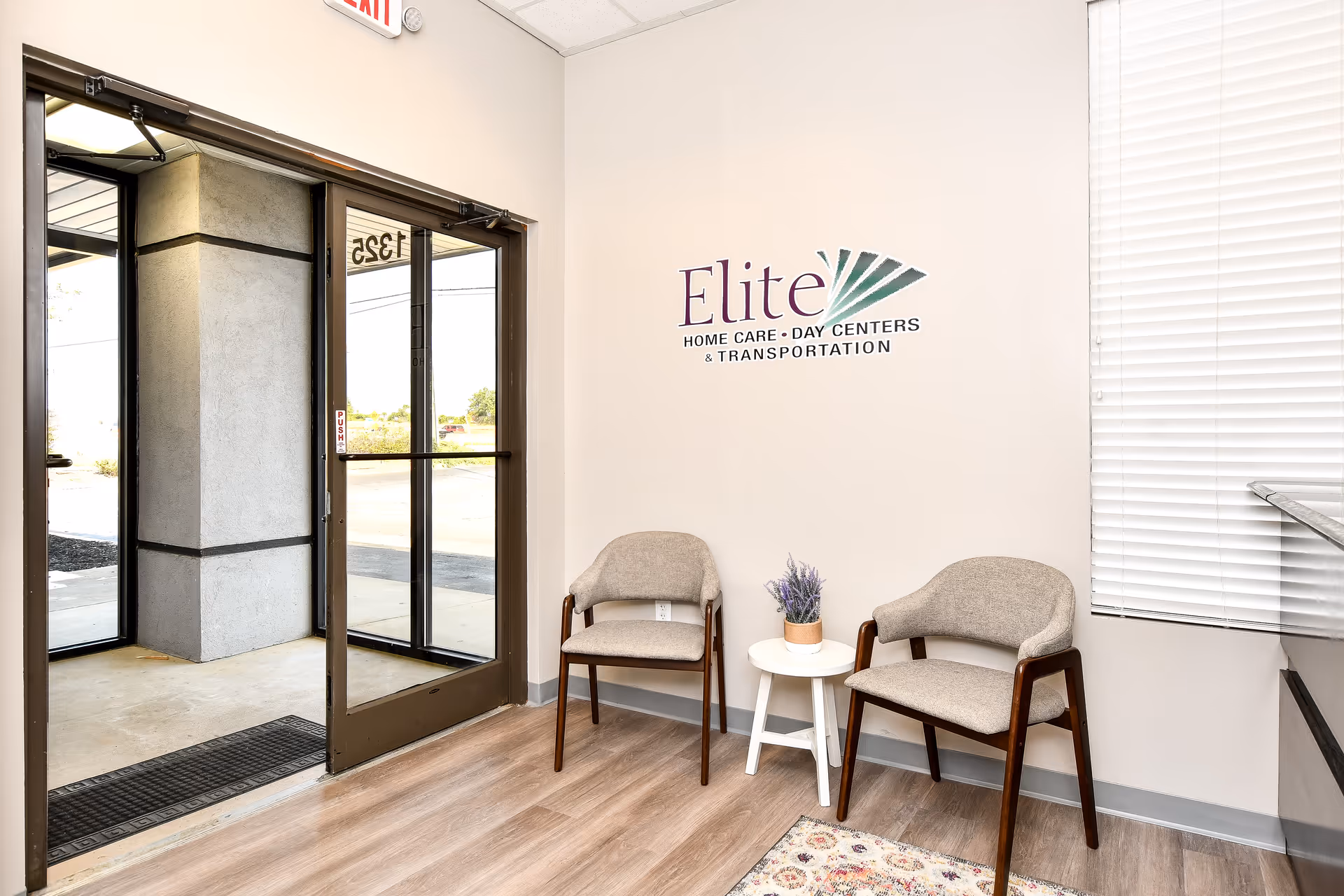 Lobby area with glass doors, two chairs, a small table with a plant, and Elite logo.