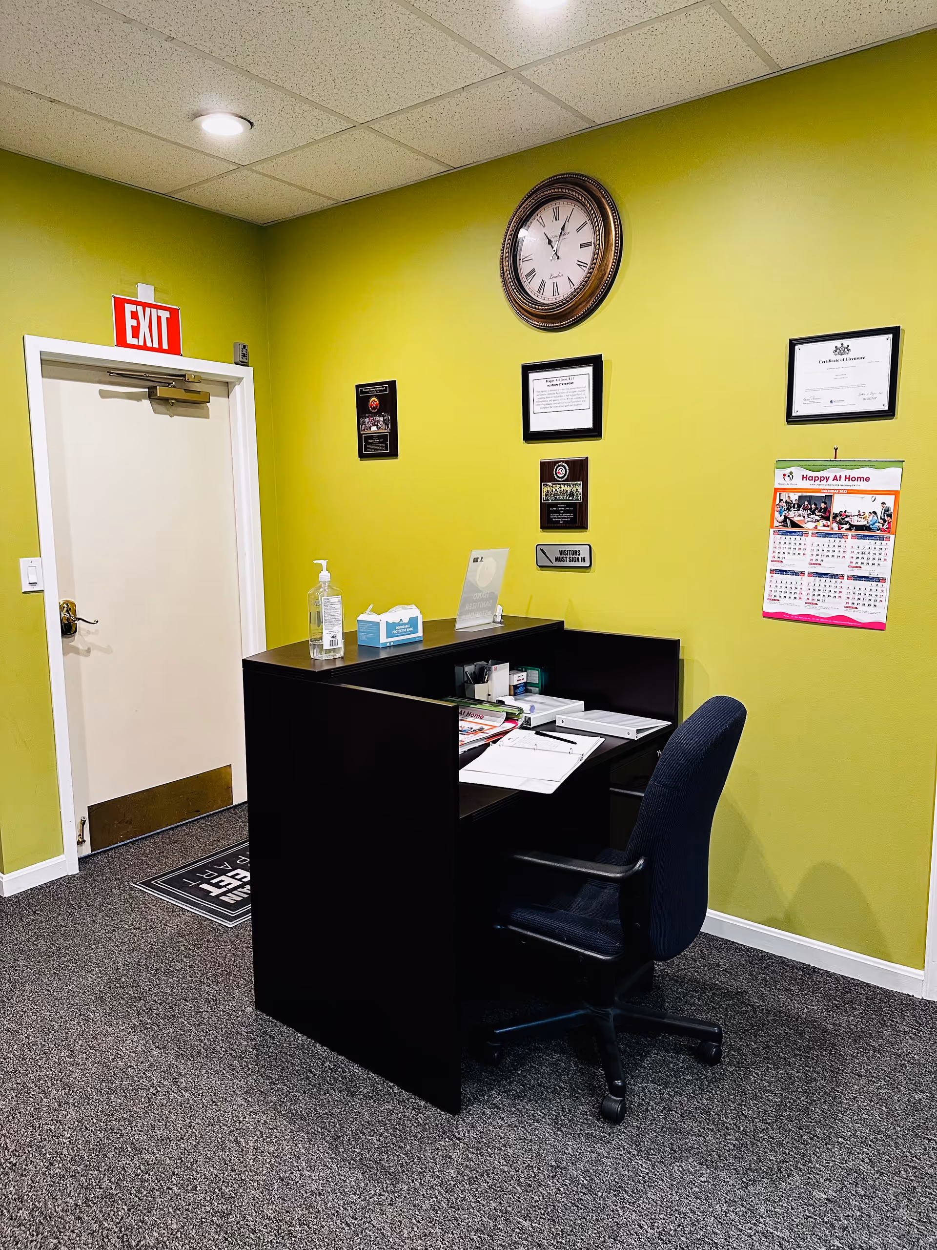 Reception desk in lime-green lobby with clock, certificates, sanitizer, tissue box, and exit sign.