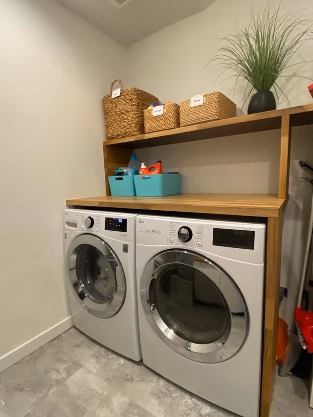 Laundry room with front-loading washing machines beneath a wooden shelf, baskets, and a plant.