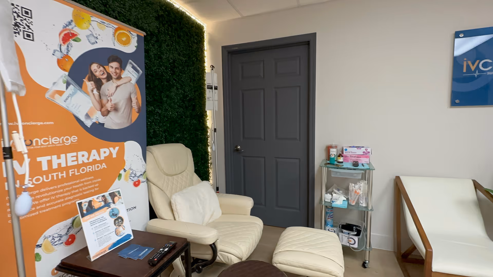 Interior reception area with beige recliner, small cart, gray door, and IV Concierge banner.