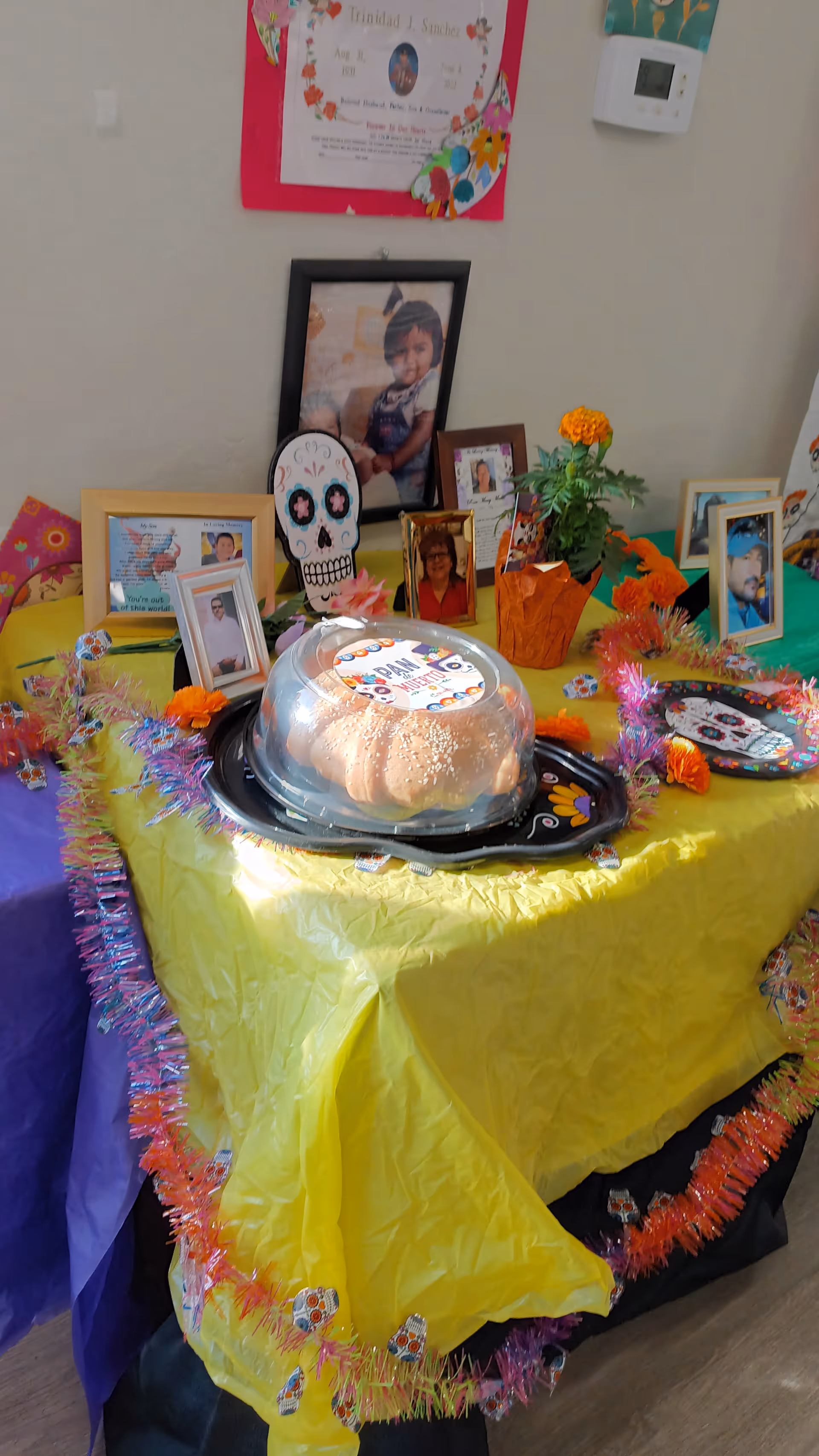 Indoor celebration setup with a cake, framed photos, flowers, and colorful table coverings.