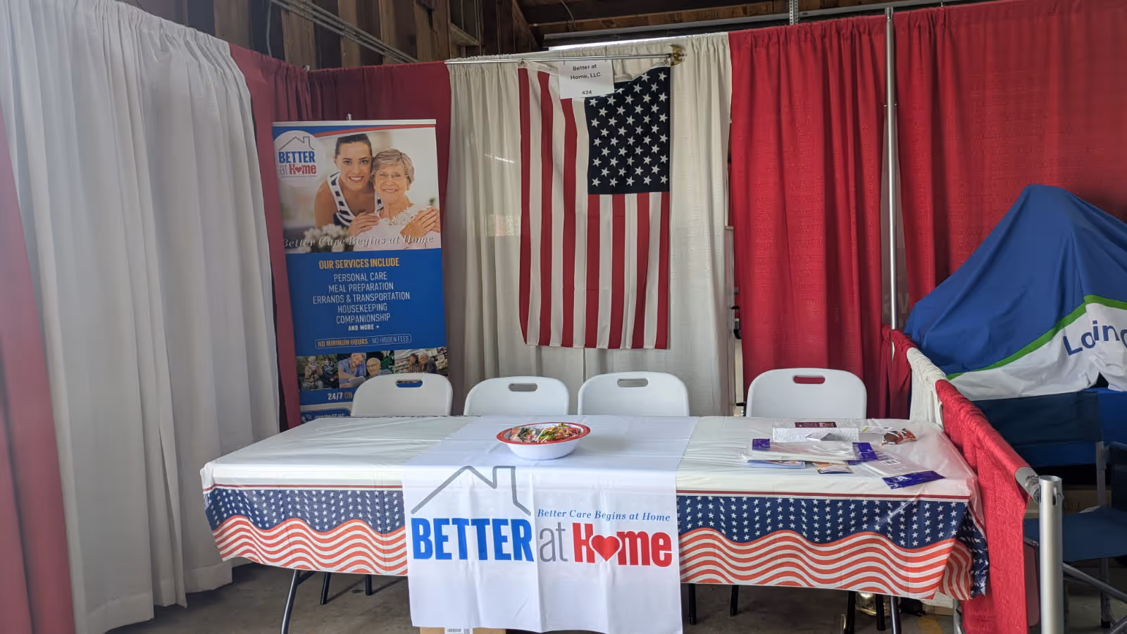 Indoor booth setup for Better At Home, LLC with banners, American flag, and a decorated table.