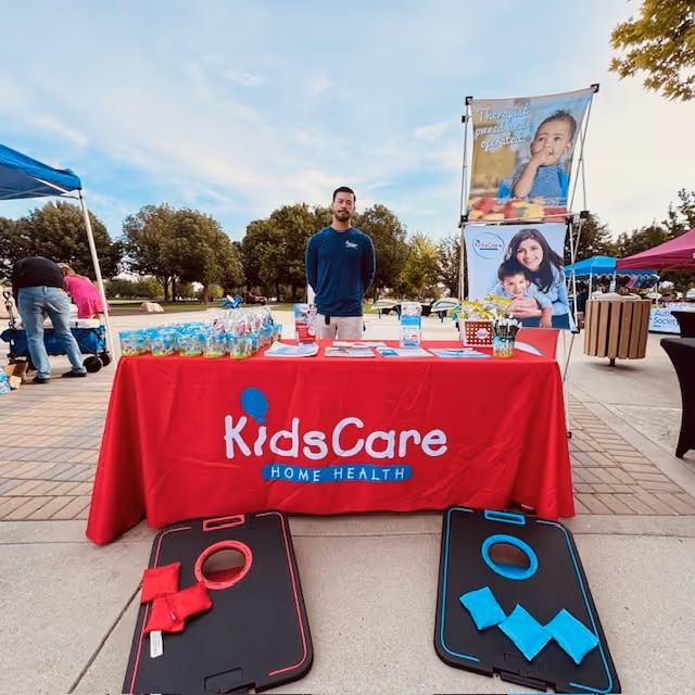 Staff member stands behind a red KidsCare Home Health table at an outdoor event.
