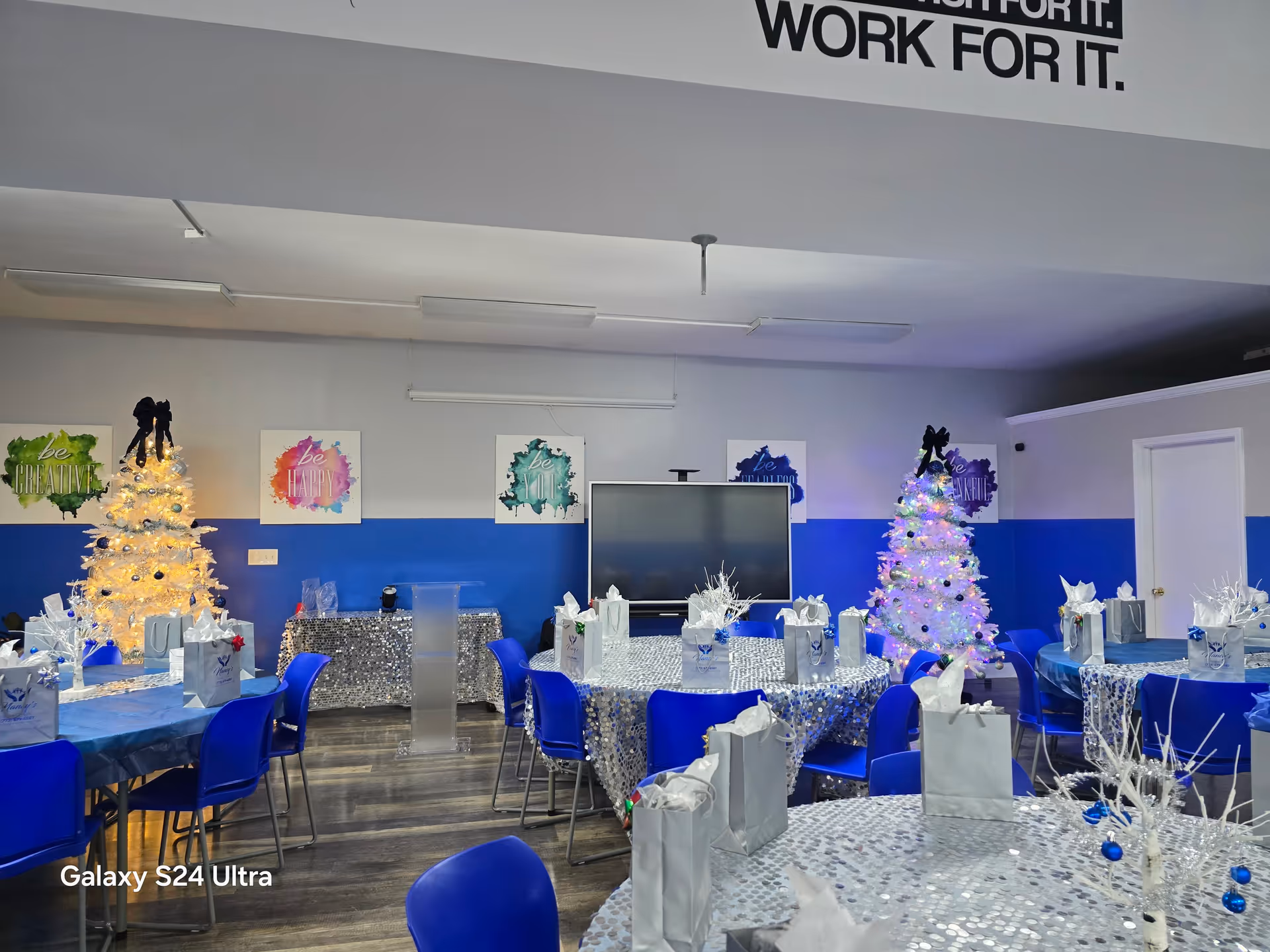 Interior event room with blue chairs, silver tables, white gift bags, and Christmas trees.