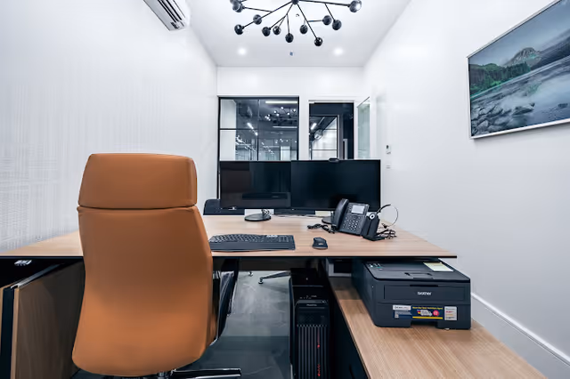 Modern office desk with dual monitors, keyboard, phone, and printer in a bright room.