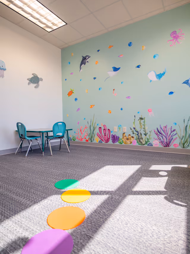 Colorful therapy room with ocean mural, teal chairs, small table, and colorful circular floor markers