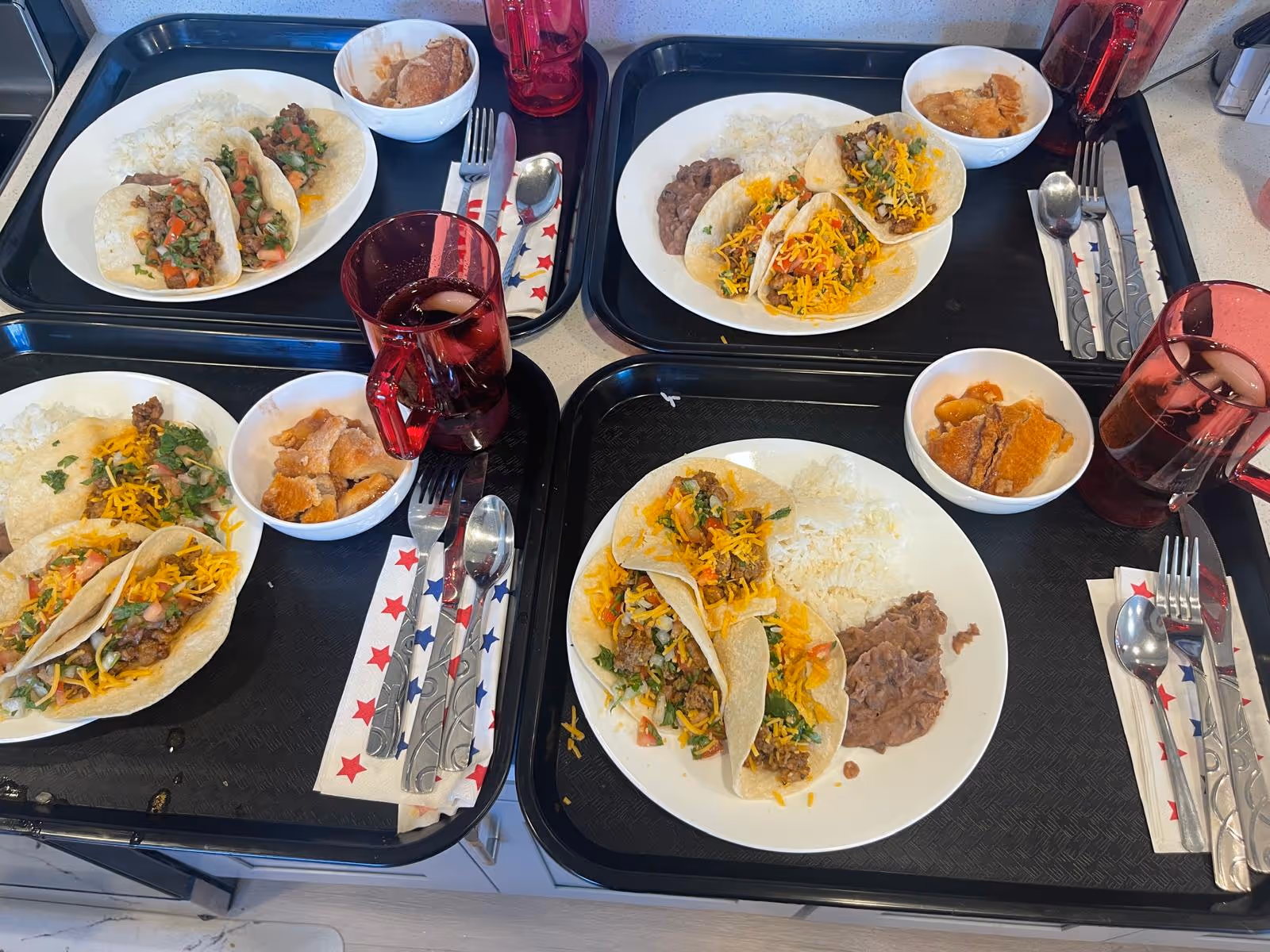Overhead view of four meal trays with tacos, rice, and beverages.