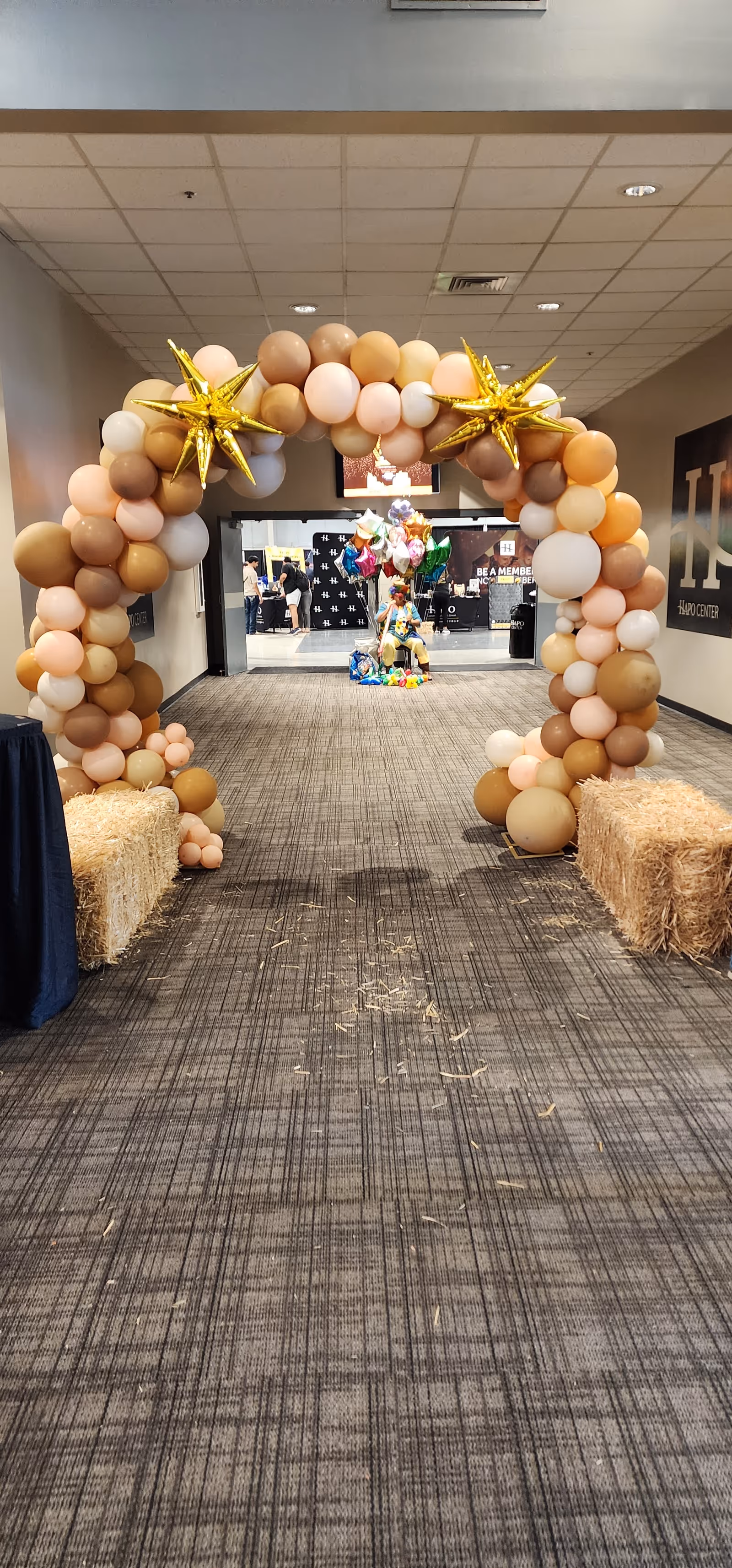 Balloon arch doorway with pastel balloons and hay bales decorates an indoor hallway.