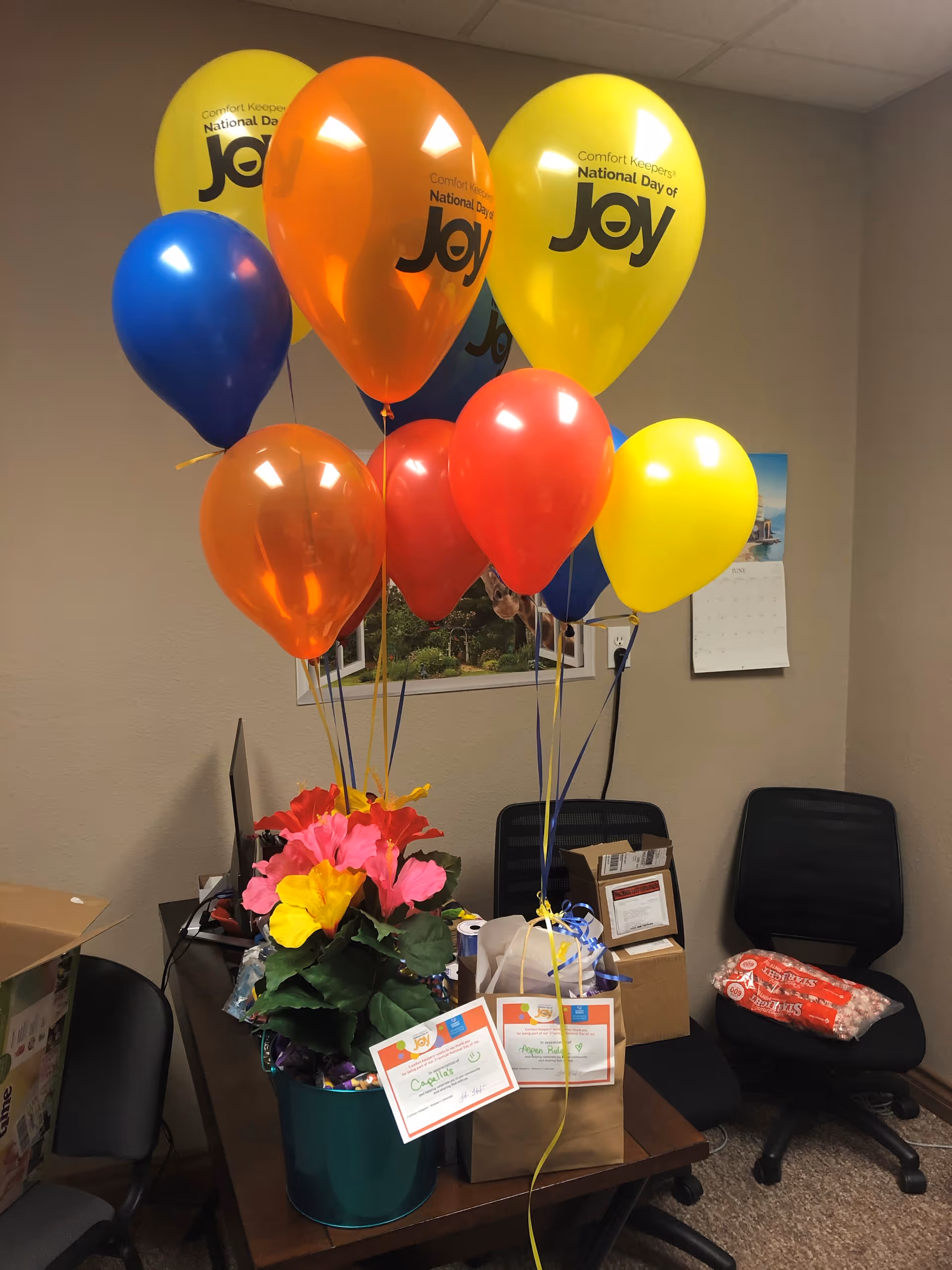Office corner with Joy balloons, flowers, gift bags, and a calendar in a Comfort Keepers room.