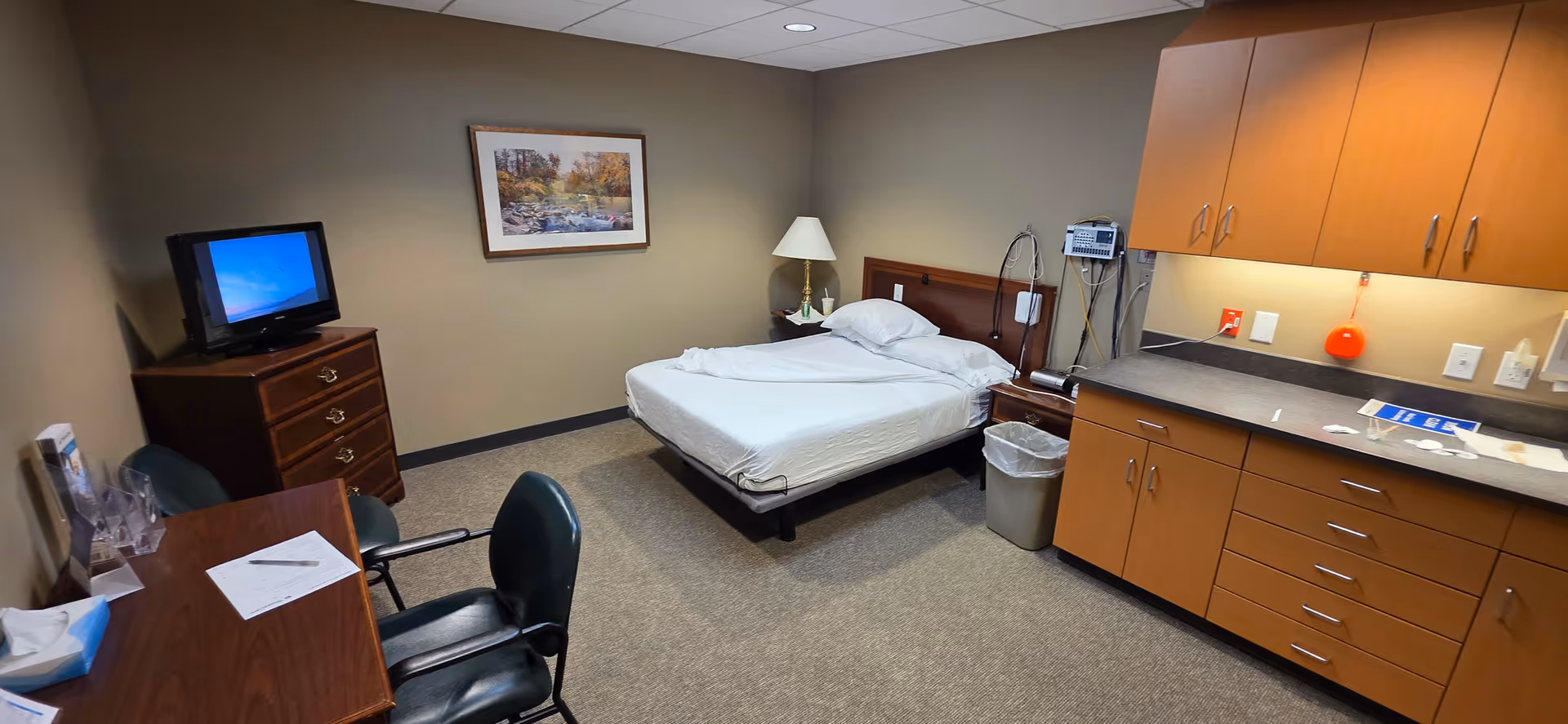 A hospital-style patient bedroom in a senior facility with a bed, desk, TV, and kitchenette area.