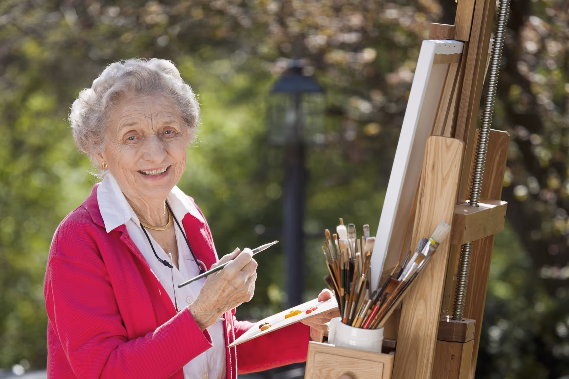 An elderly woman in a pink jacket paints outdoors at an easel, smiling.