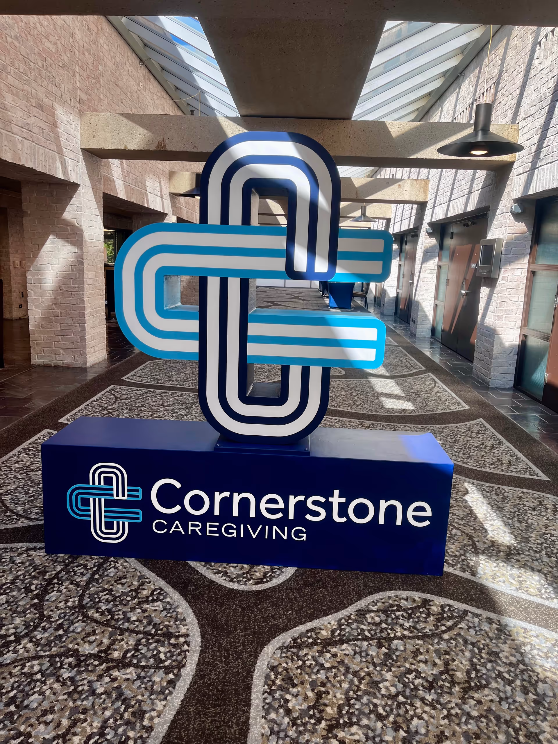 Indoor atrium corridor with a large Cornerstone Caregiving sign sculpture and patterned floor.