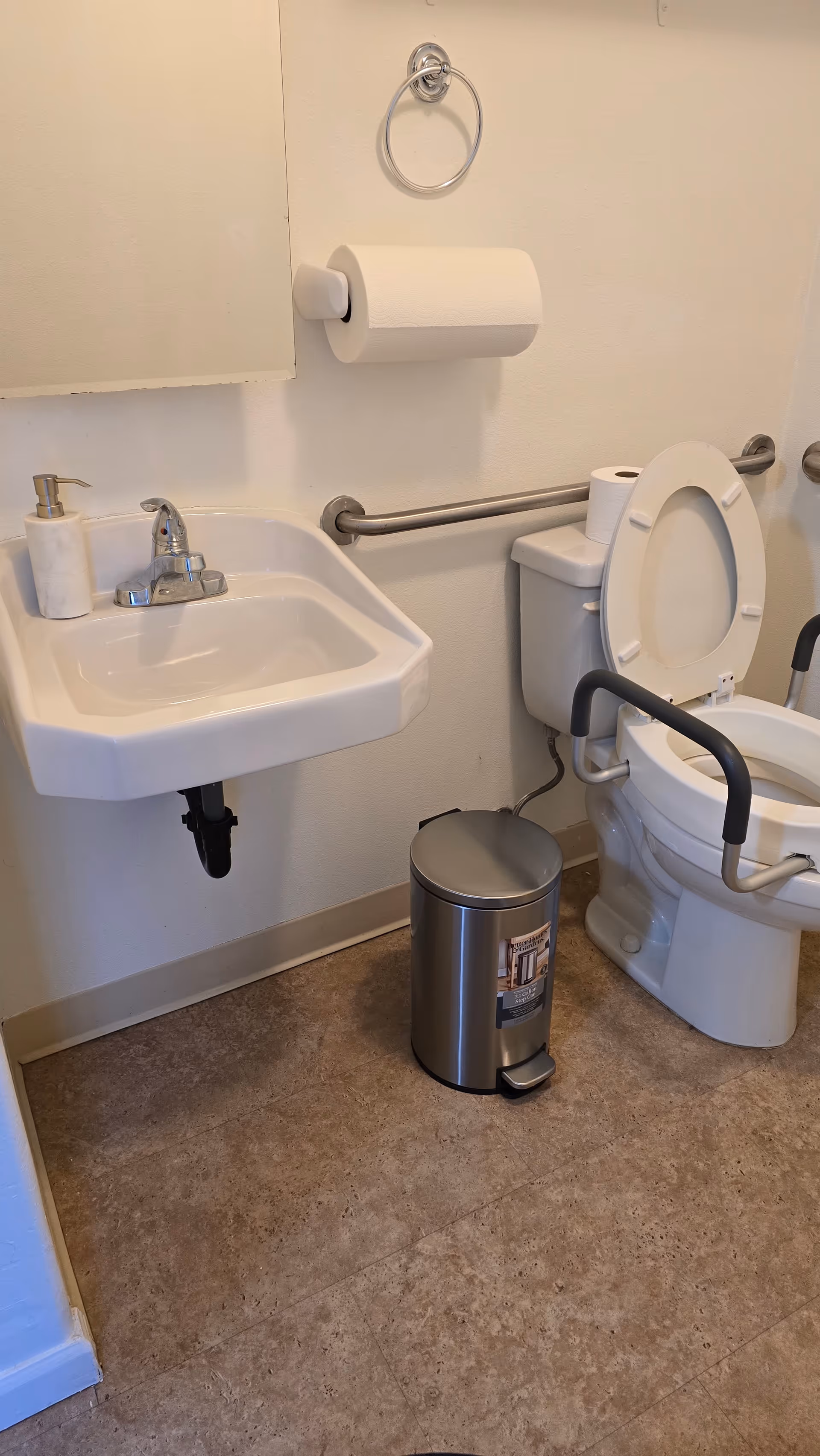 Small bathroom with sink, toilet, grab bars, toilet paper, and a trash can.