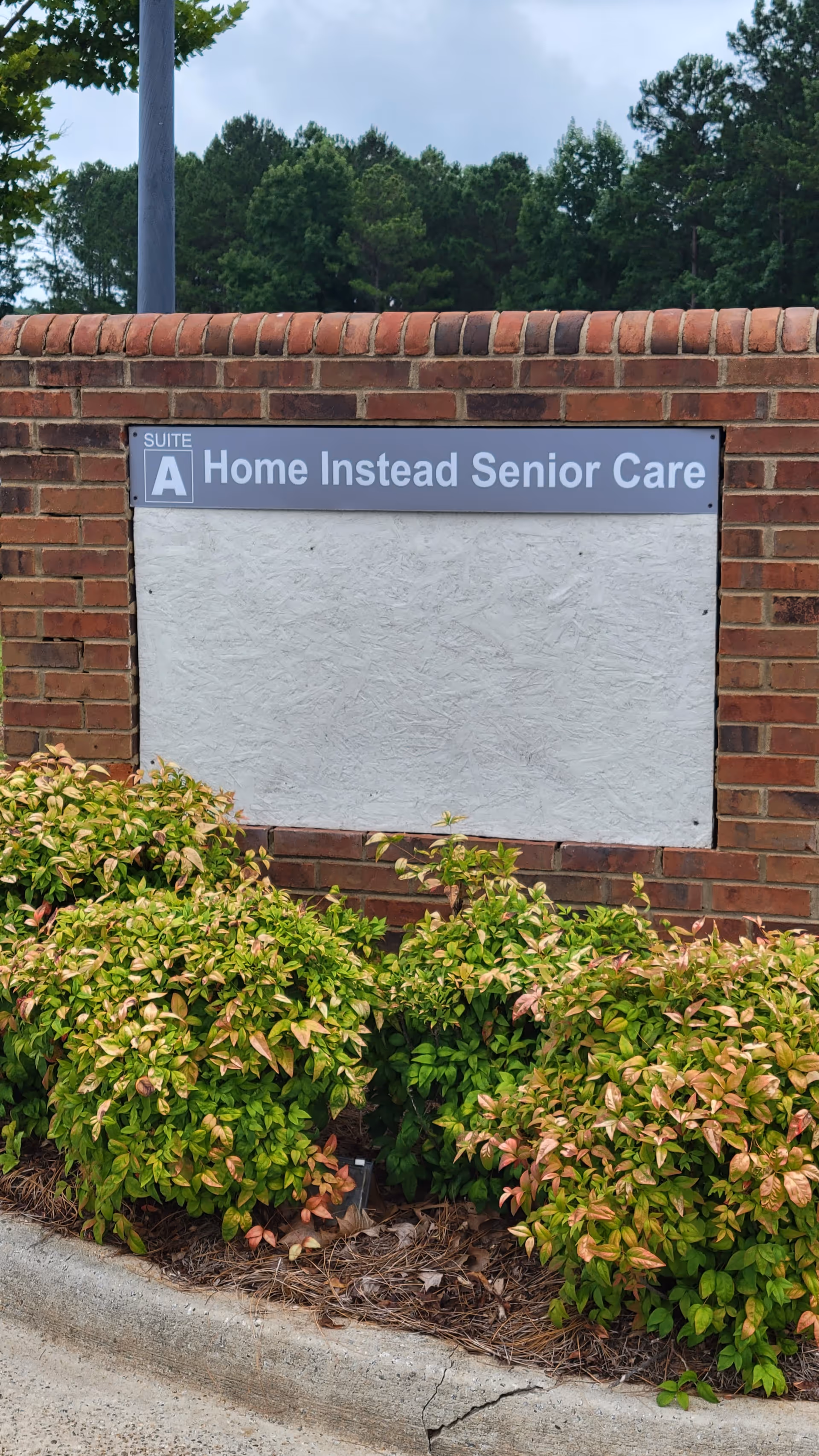 Exterior brick wall with a sign reading Home Instead Senior Care and surrounding shrubbery.