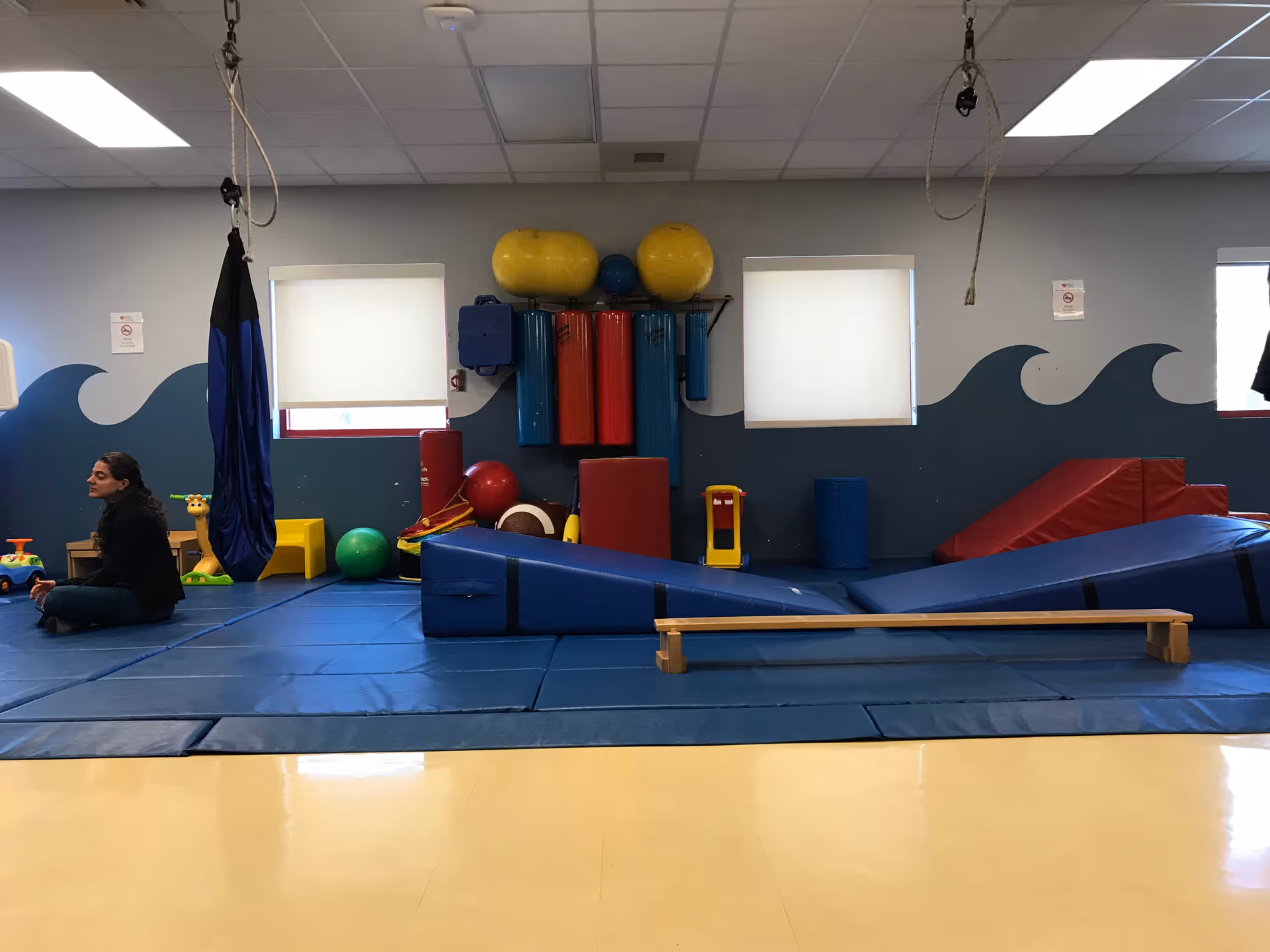 Indoor play area with blue mats, climbing blocks, and a seated person near equipment.